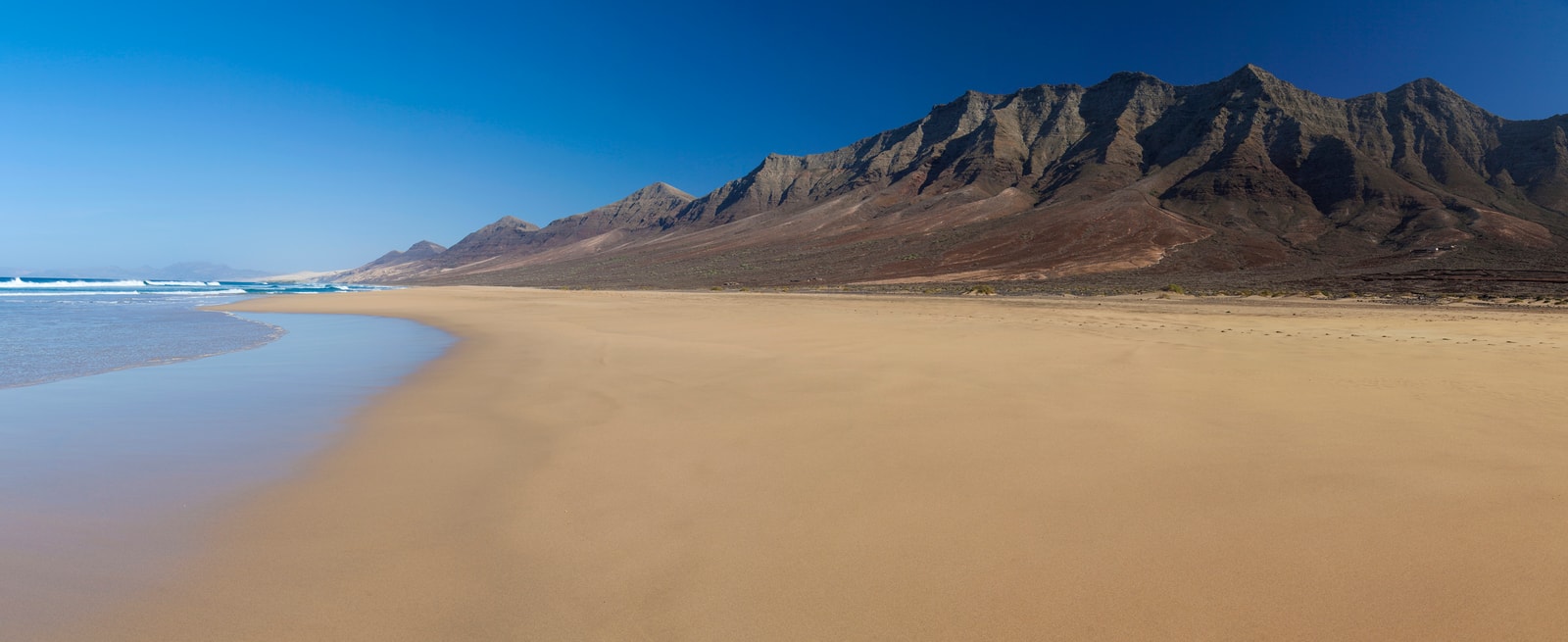 A panoramic ground-level view of Cofete beach shows the sky reflecting on wet golden sand, with massive mountains rising in the background.