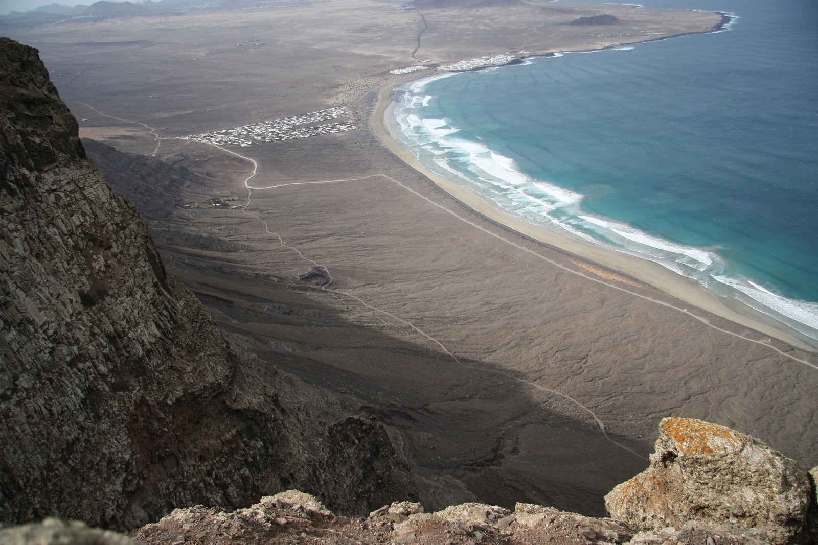 A high-angle view shows a long, curving beach at the base of massive, rugged cliffs with a small town nearby.