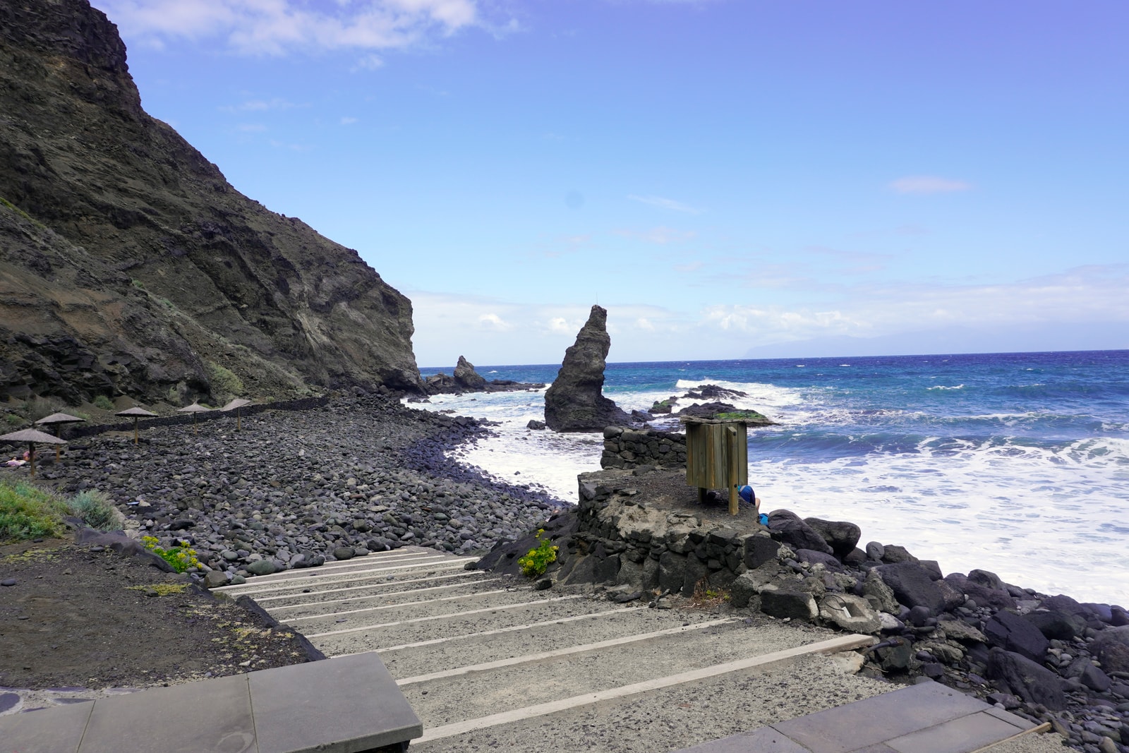 A remote black-pebble cove at Playa de la Caleta, with a sea stack, dramatic cliffs, and visible access steps.