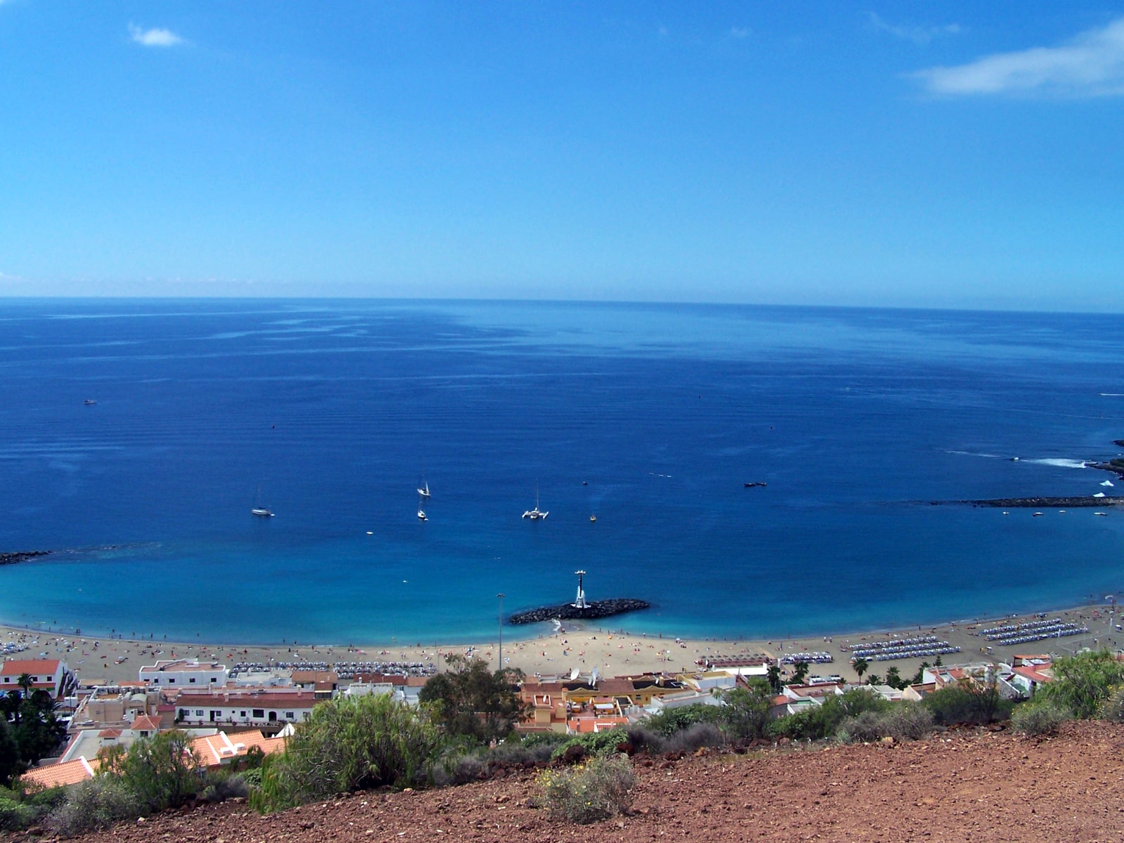 An elevated view shows a crescent-shaped beach with blue water, dotted with umbrellas and boats, next to a harbor breakwater.