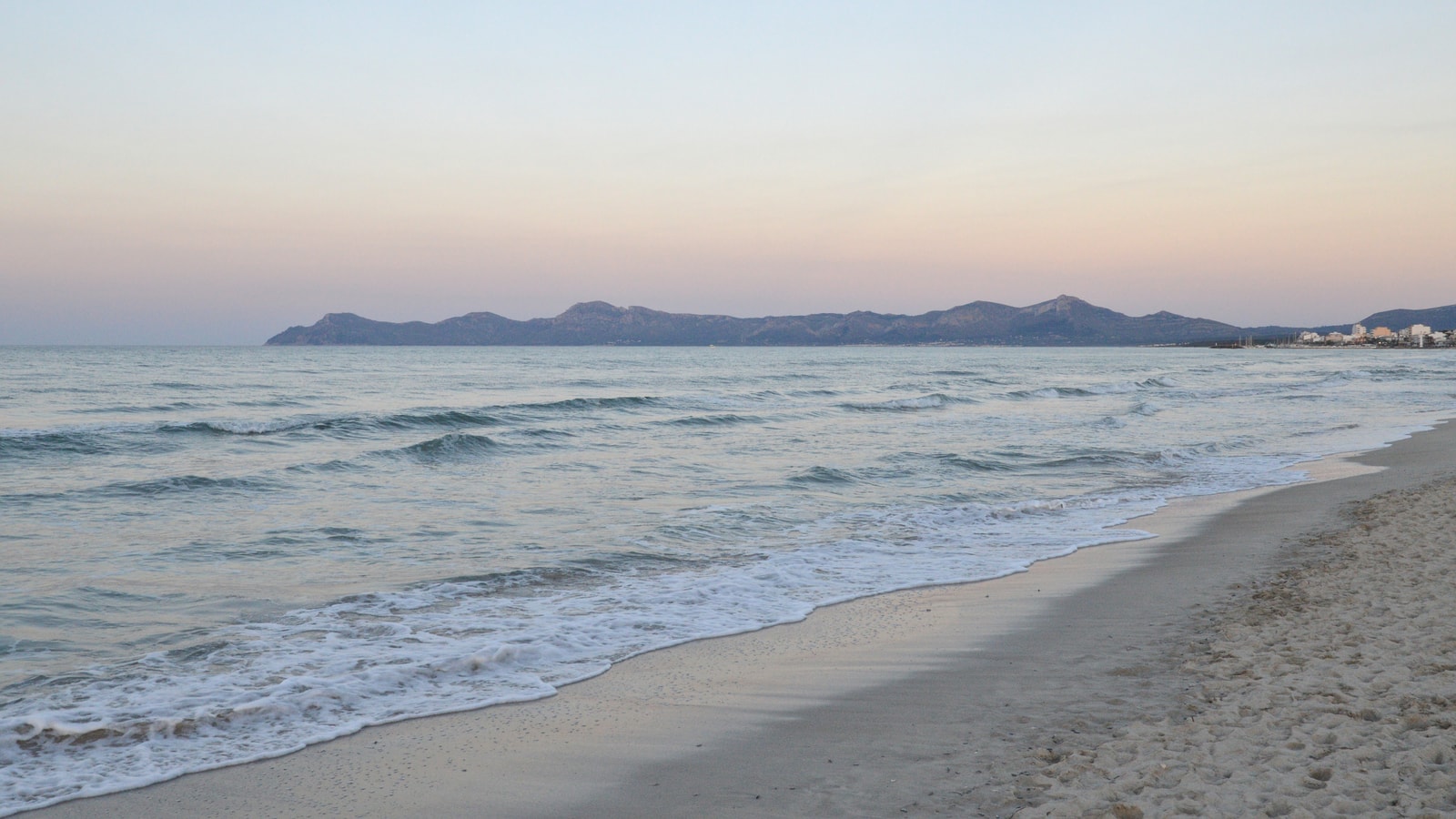 Calm, gentle waves lap onto a wide, empty sandy beach under a partly cloudy sky, with a distant headland on the horizon.