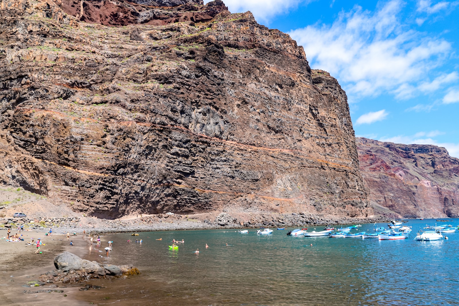 Layered cliff walls rise above the sheltered water of Playa de Vueltas, where boats are moored and people swim.
