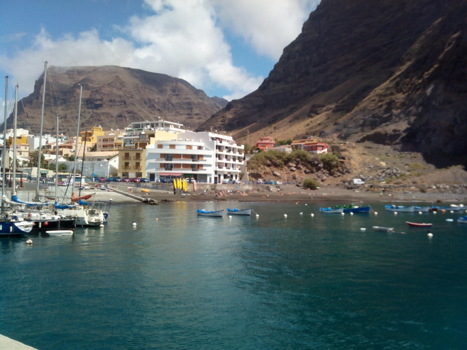 A view of the sheltered harbor at Playa de Vueltas, with moored boats, waterfront buildings, and steep slopes.