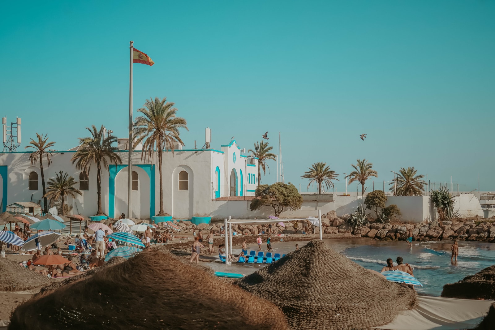 The bustling urban shore of Playa del Faro in Marbella, with its signature thatched umbrellas, golden sand, and city buildings in the background.