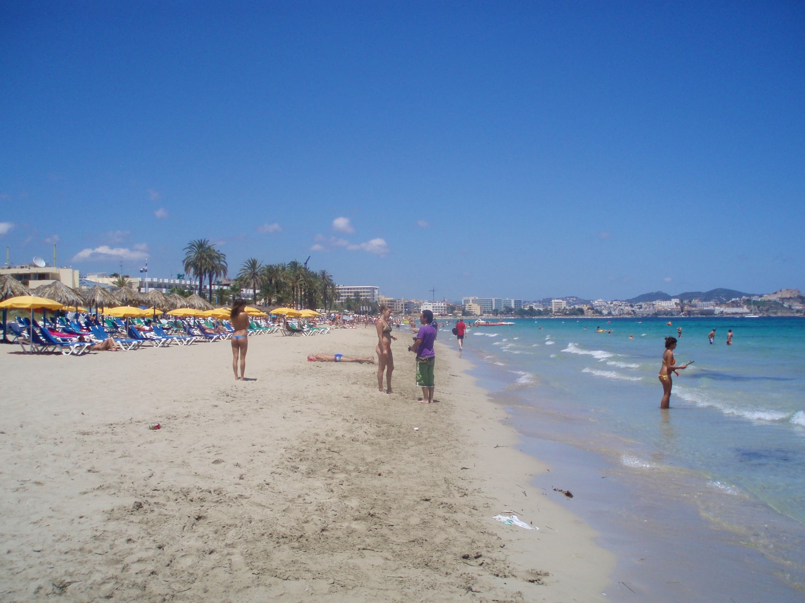 A bustling daytime scene at Playa d'en Bossa, with yellow parasols and sun loungers in front of the Bora Bora club.