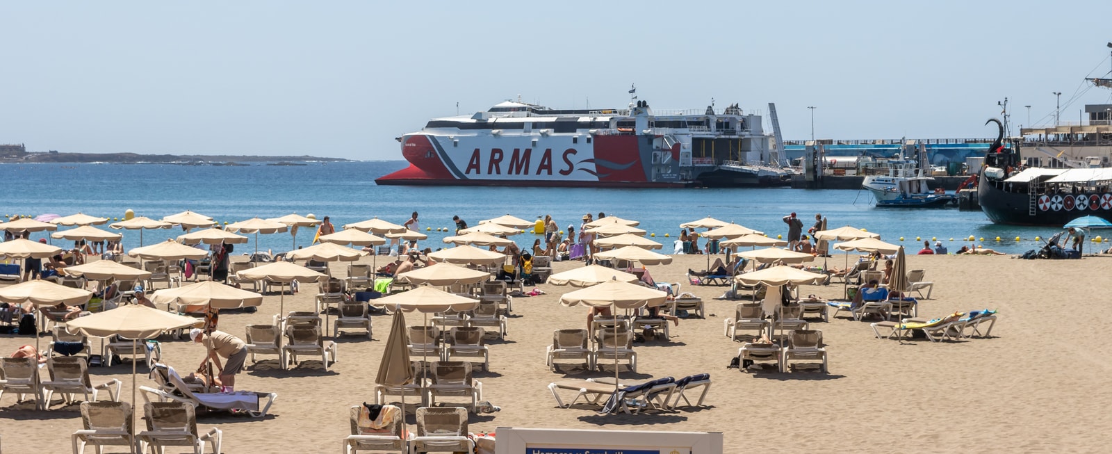 A wide view of a beach with parasols, with a large white ferry docked in the harbor behind it.