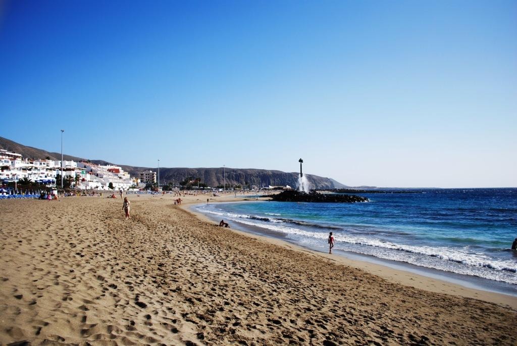 Textured sand in the foreground leads to gentle blue waves breaking on the shore, with a rocky breakwater in the distance.