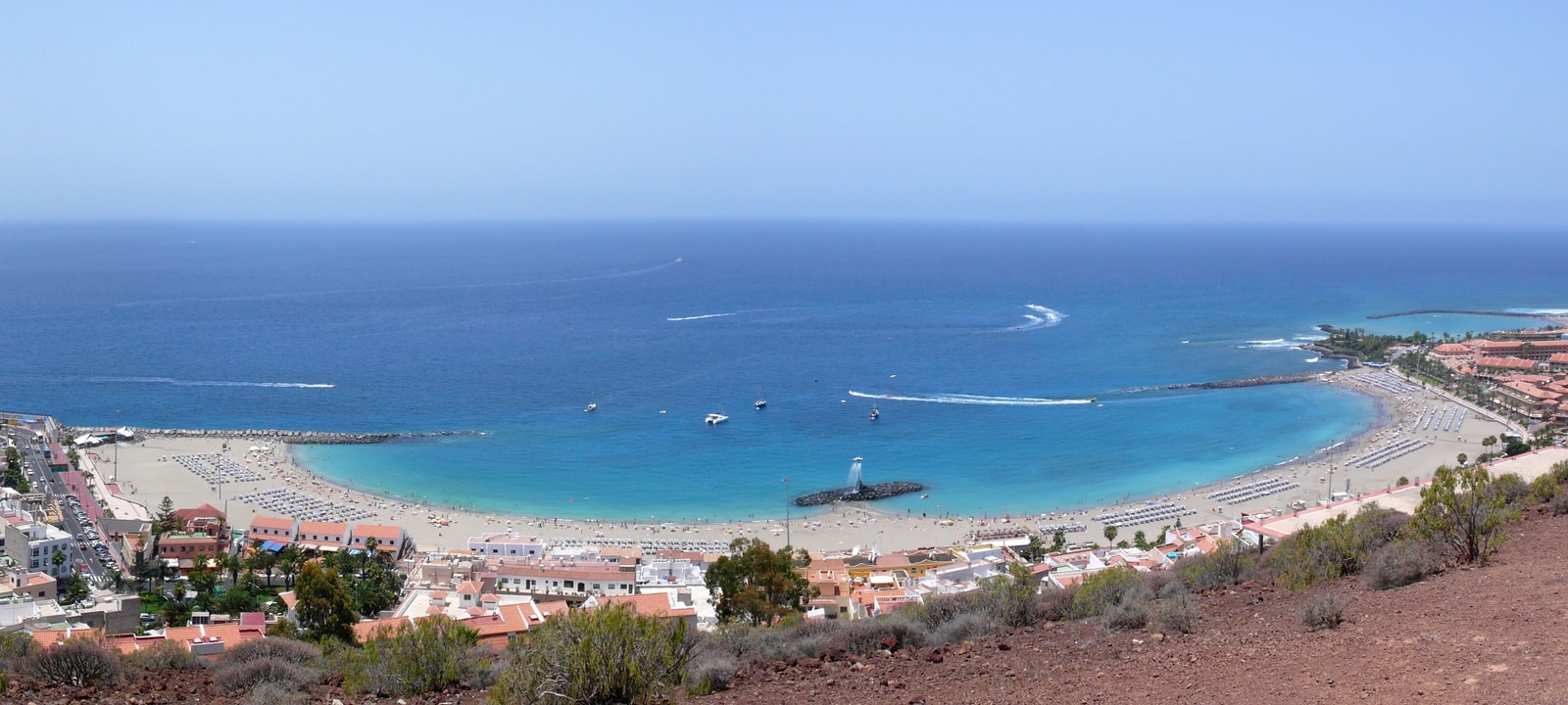 A wide panoramic view of a large, curved sandy beach packed with visitors, with a resort town and breakwaters enclosing the bay.