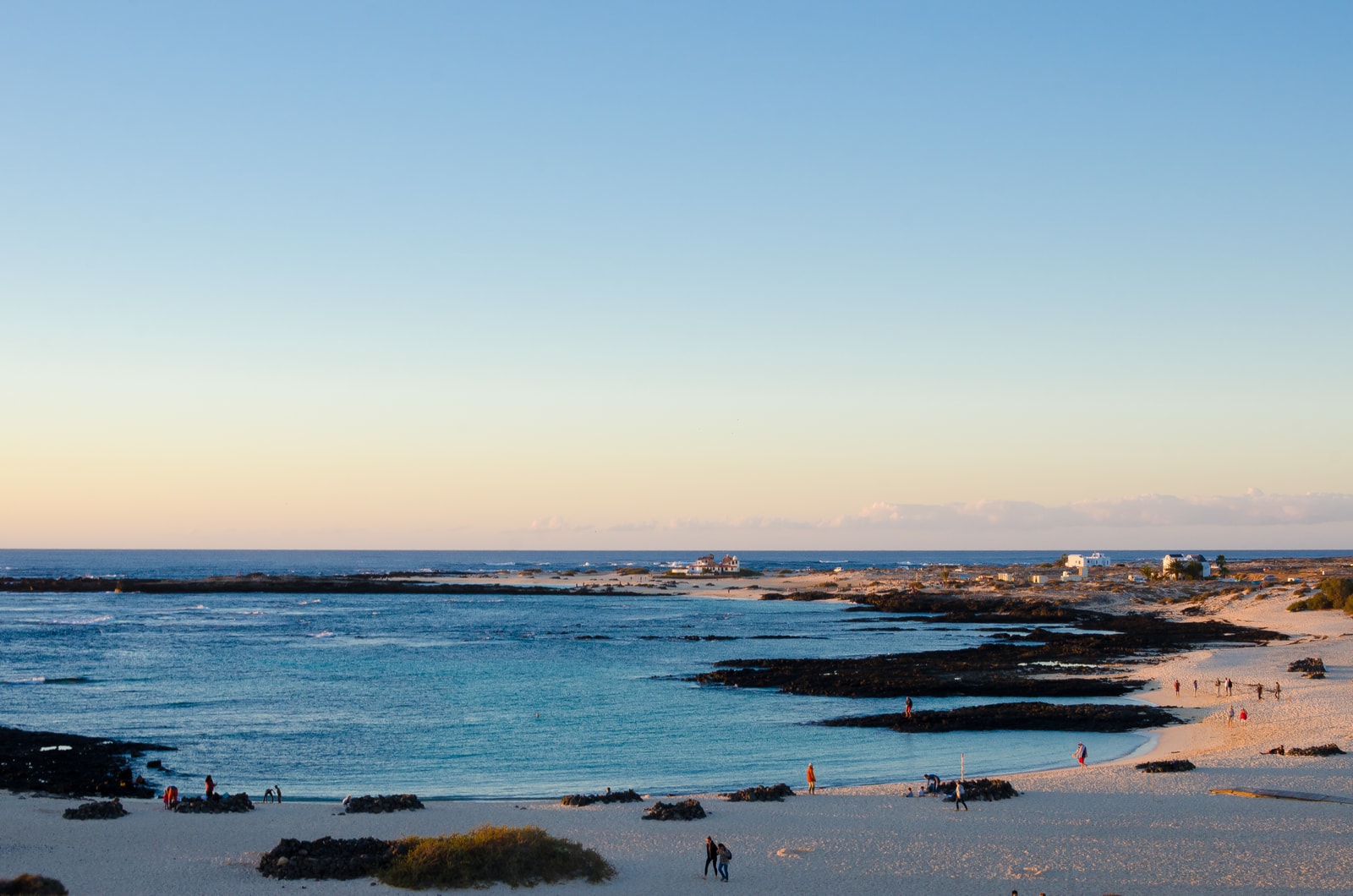 A beach with calm, shallow pools of water formed by dark volcanic reef shelves, with people relaxing on the white sand.