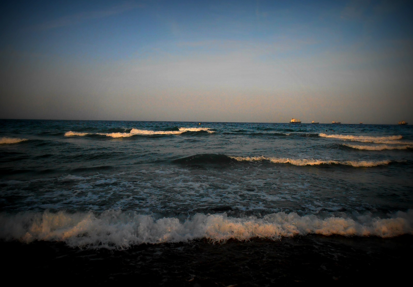 Waves wash onto the dark sand of Malvarrosa beach at dusk, with cargo ships visible on the horizon.