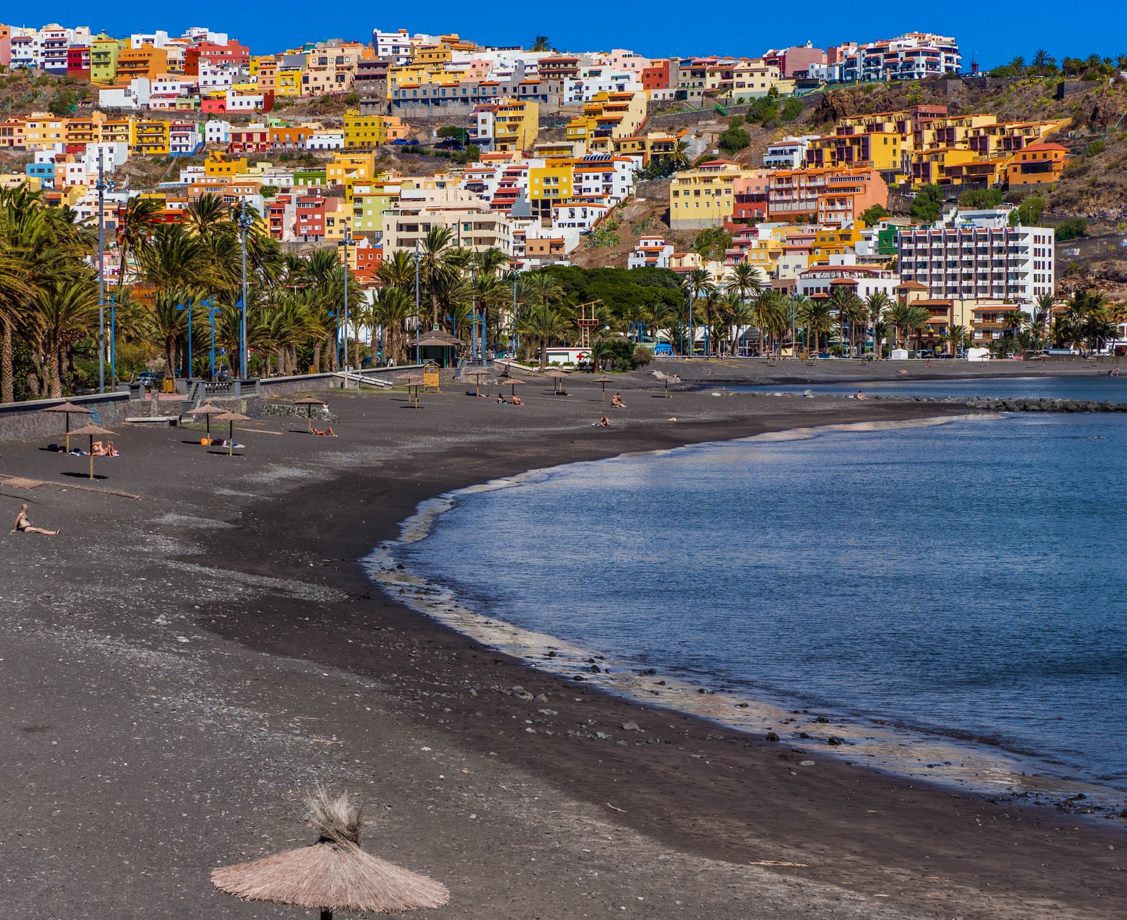 A view of the dark sand beach and palm-lined promenade in San Sebastián de La Gomera.