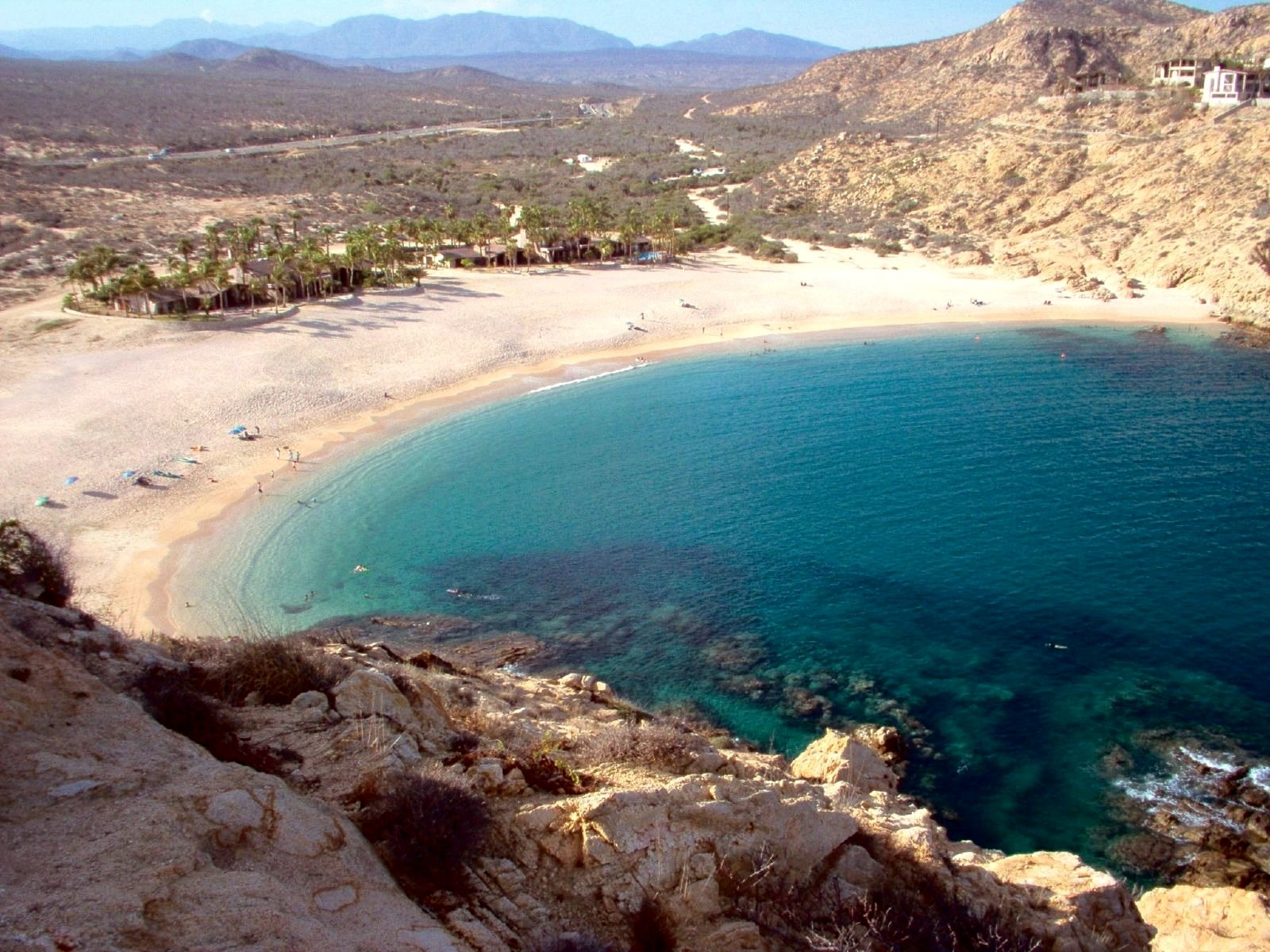 An elevated view of Playa Santa Maria's curved, sandy cove, with turquoise water and the surrounding rocky, arid landscape of Baja California.