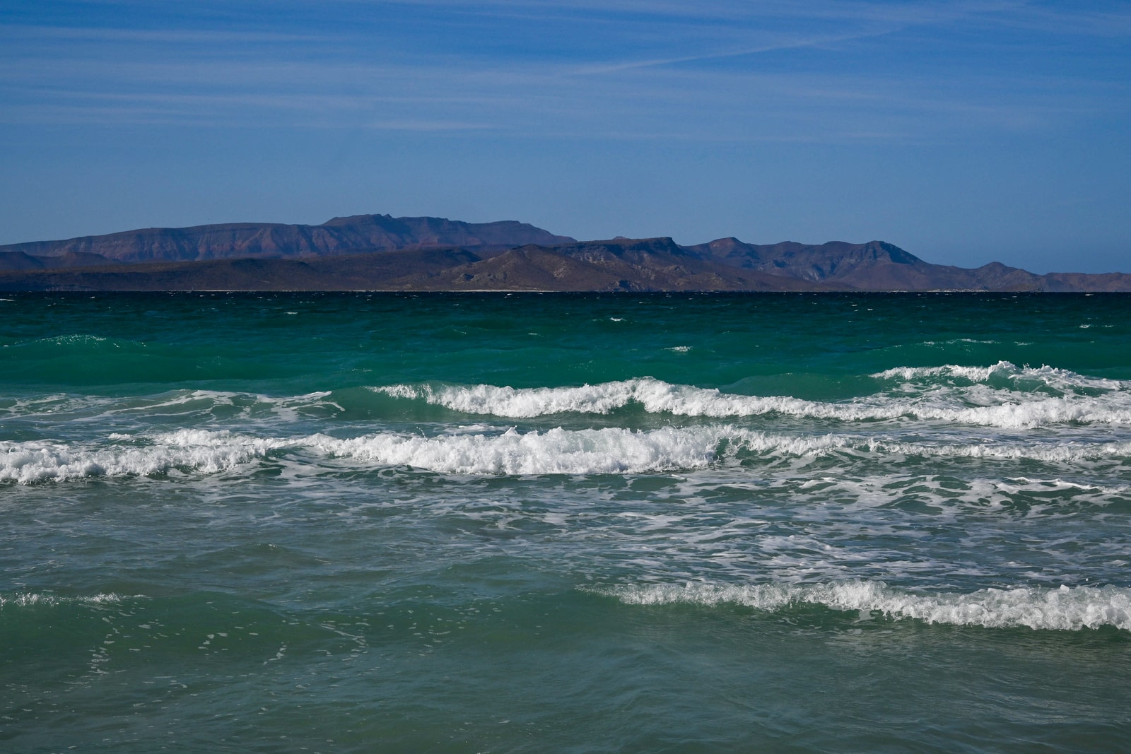A view of Playa Tecolote, with gentle waves on the shore, a wide sandy beach, and rugged mountains in the distance.