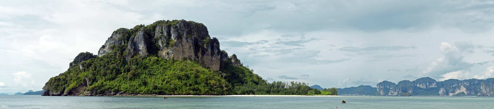 A panoramic view of Poda Island shows a long stretch of white sand, calm turquoise water, and dramatic limestone karst formations under a blue sky.
