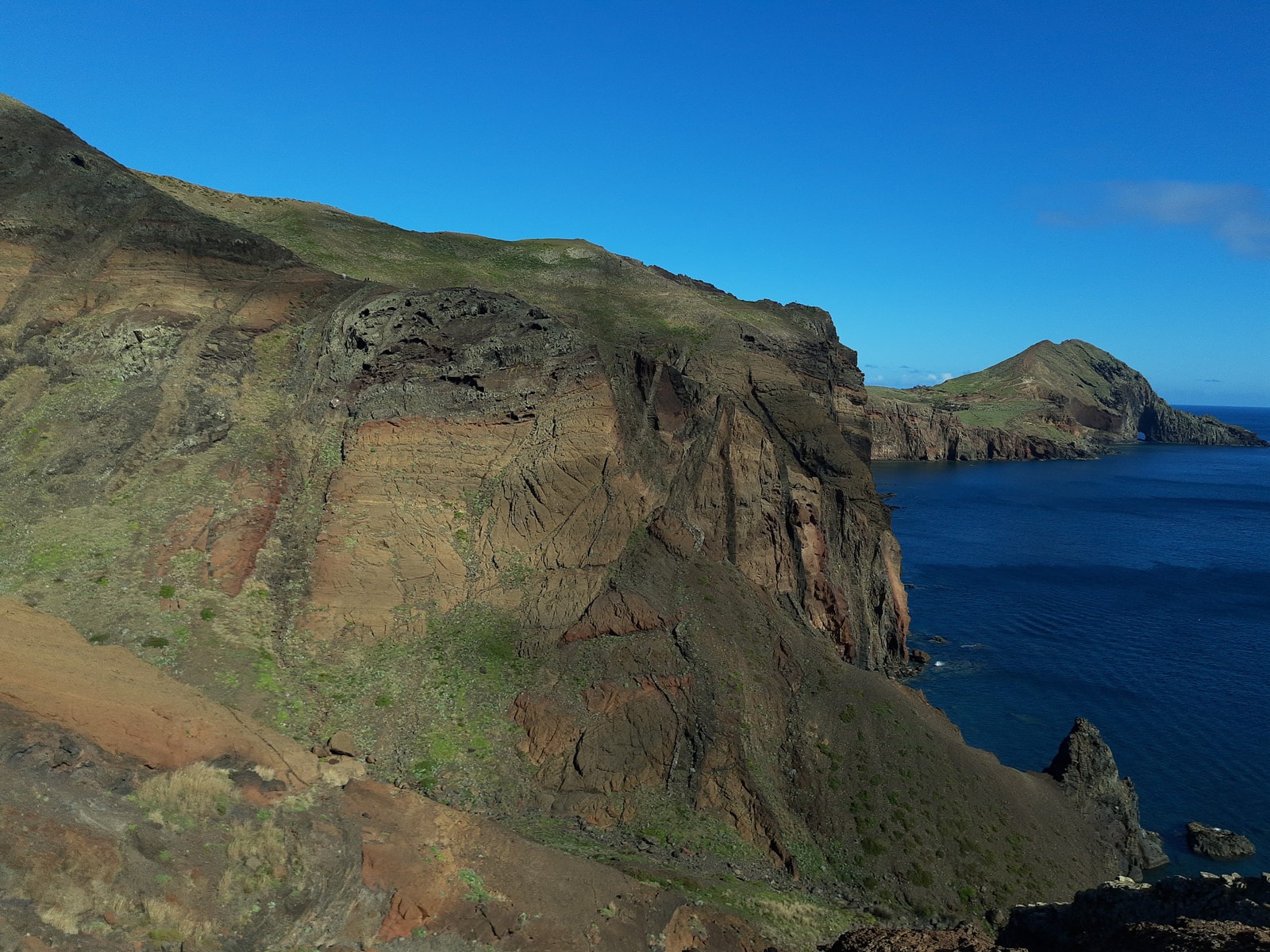 A secluded beach with dark, reddish-brown sand nestled between dramatic, stratified rock formations.