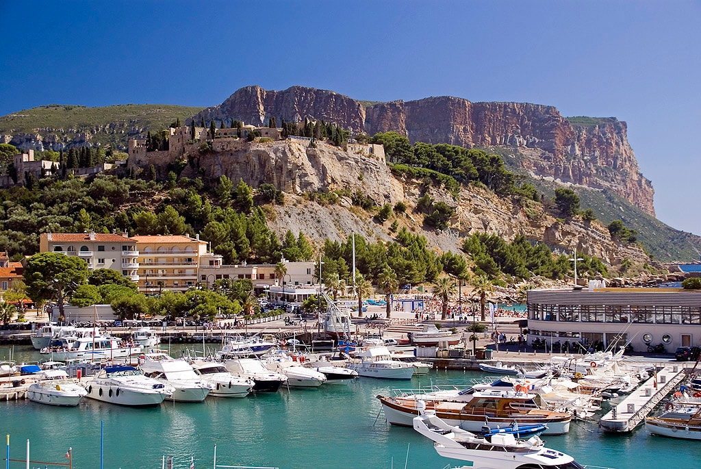 A landscape photo of the Port de Cassis, showing docked yachts, the town on the hillside, and the massive sea cliff of Cap Canaille in the background.