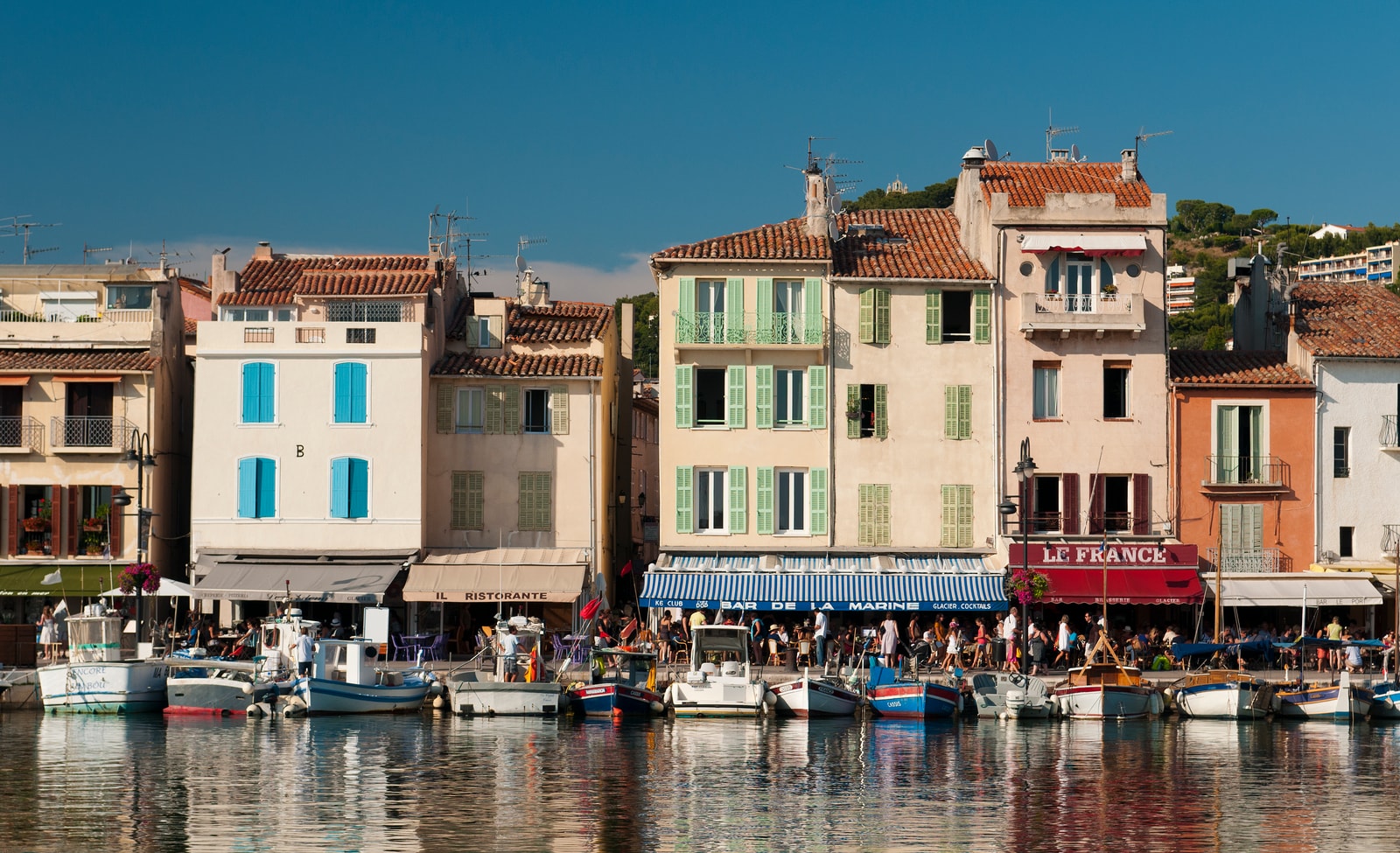 The harbor of Cassis, with pastel-colored buildings lining the waterfront, small fishing boats, and the large Cap Canaille cliff in the background under a blue sky.