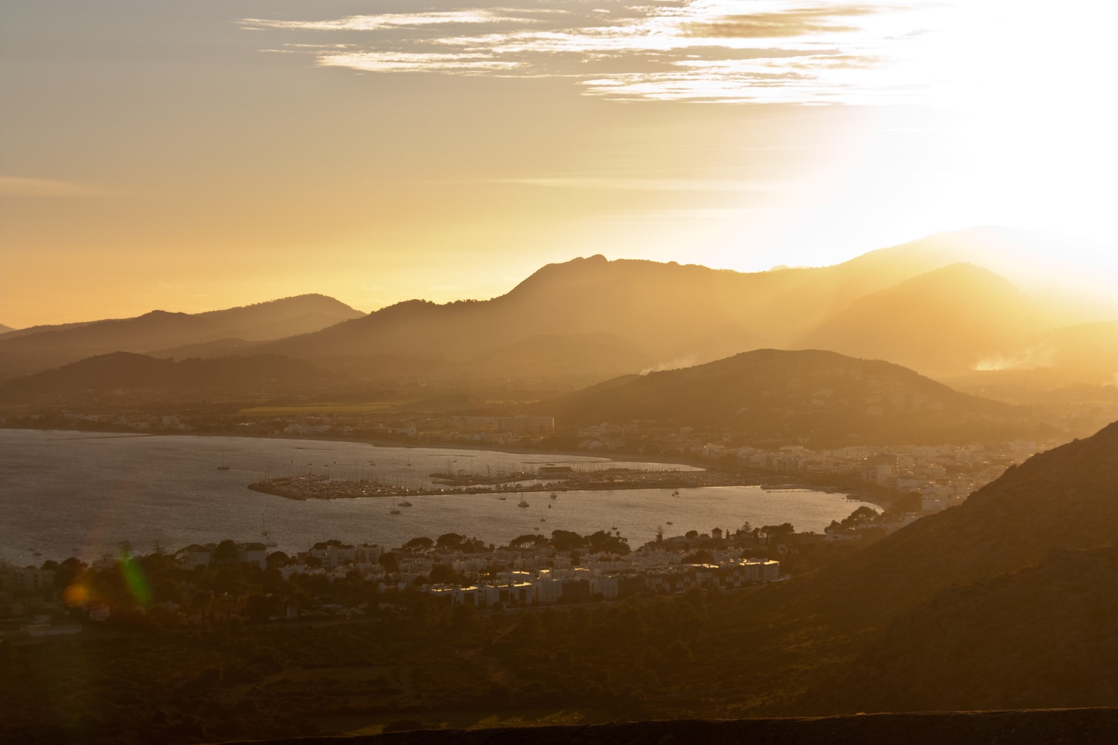 A golden sunset reflects on the calm water of a bay, with silhouetted mountains in the background and boats moored near the shore.