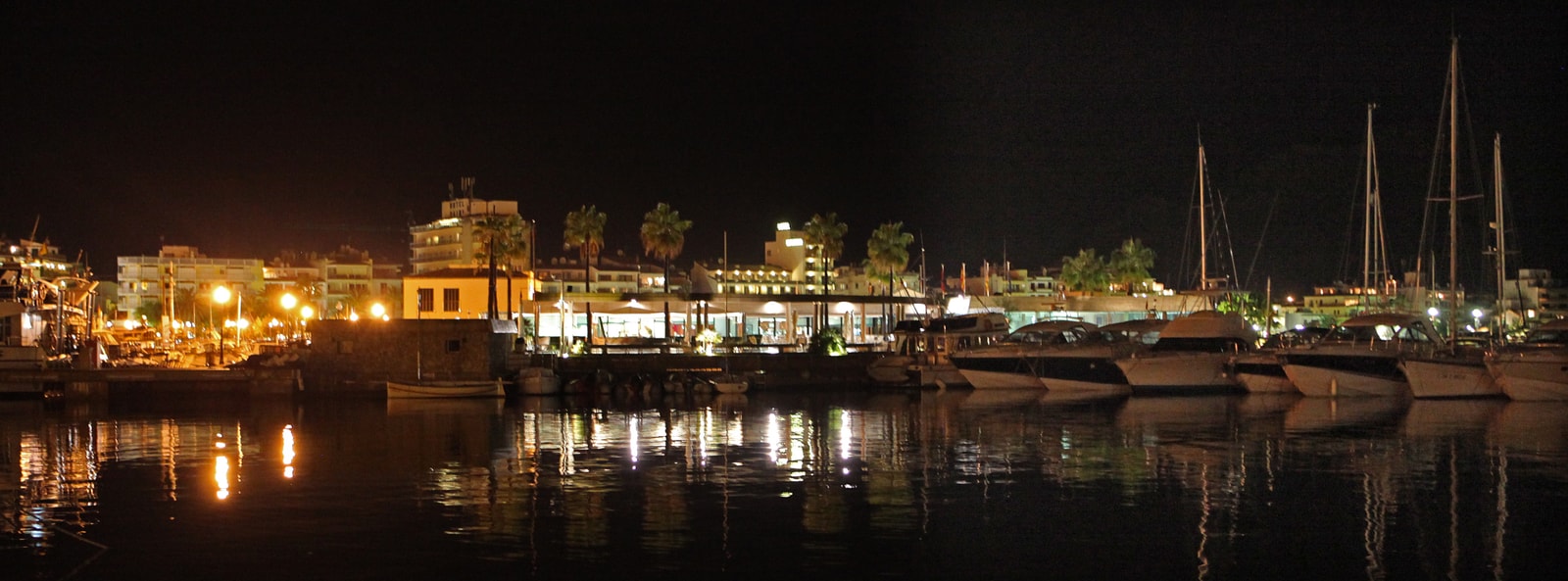 A panoramic view of a marina at night, with boats, illuminated buildings, and palm trees reflected in the dark water.