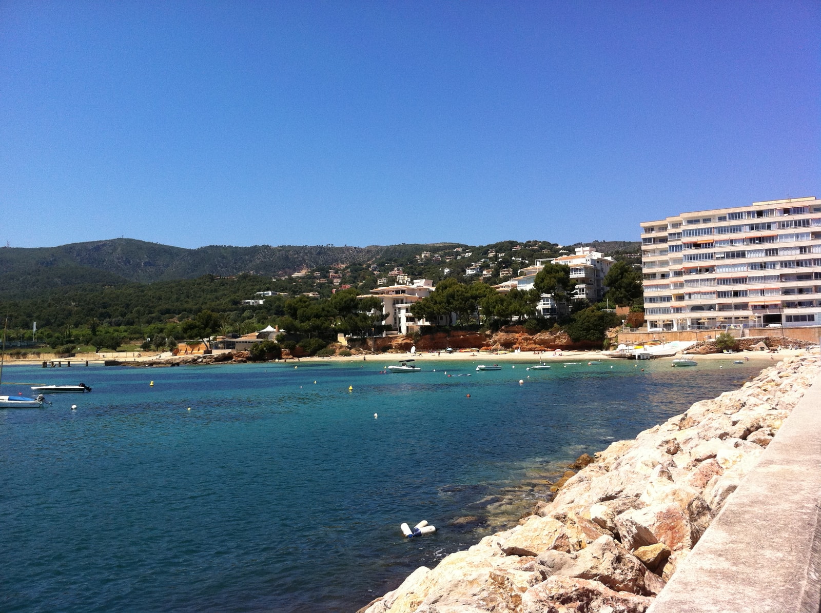 A sandy beach with clear blue water, with boats in the background and developed shoreline featuring large hotel buildings.