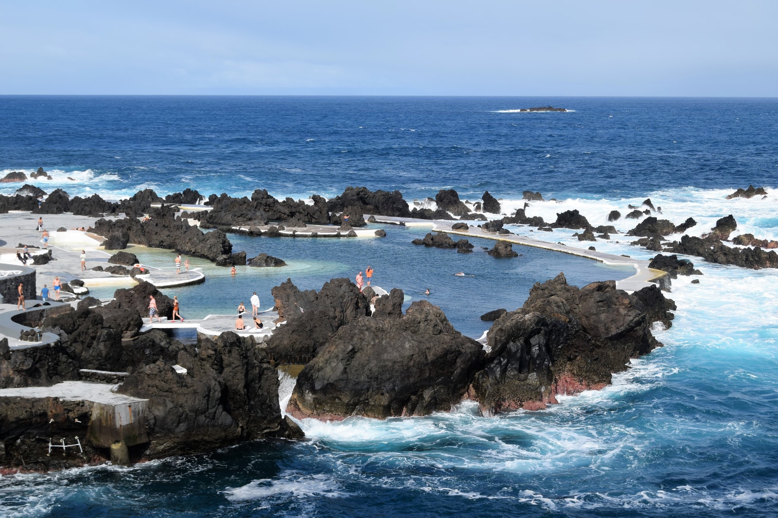 Turquoise water fills natural swimming pools formed by dark volcanic rock, with the open ocean visible beyond.