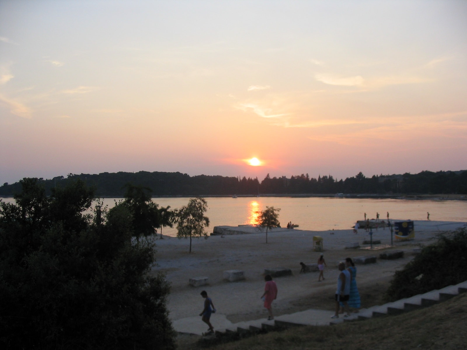 Bathers enjoy a warm sunset at Porton Biondi, with stone steps leading into the water and a gravel shore.