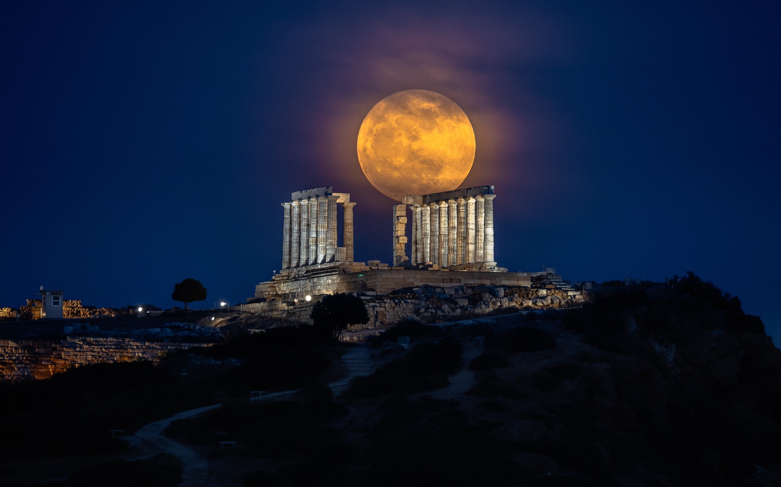 A dramatic photo of the illuminated Temple of Poseidon at night with a large full moon positioned directly behind it.