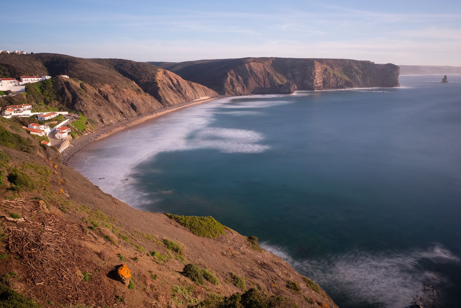 A panoramic view of a crescent-shaped bay surrounded by steep cliffs with waves breaking on the shore.