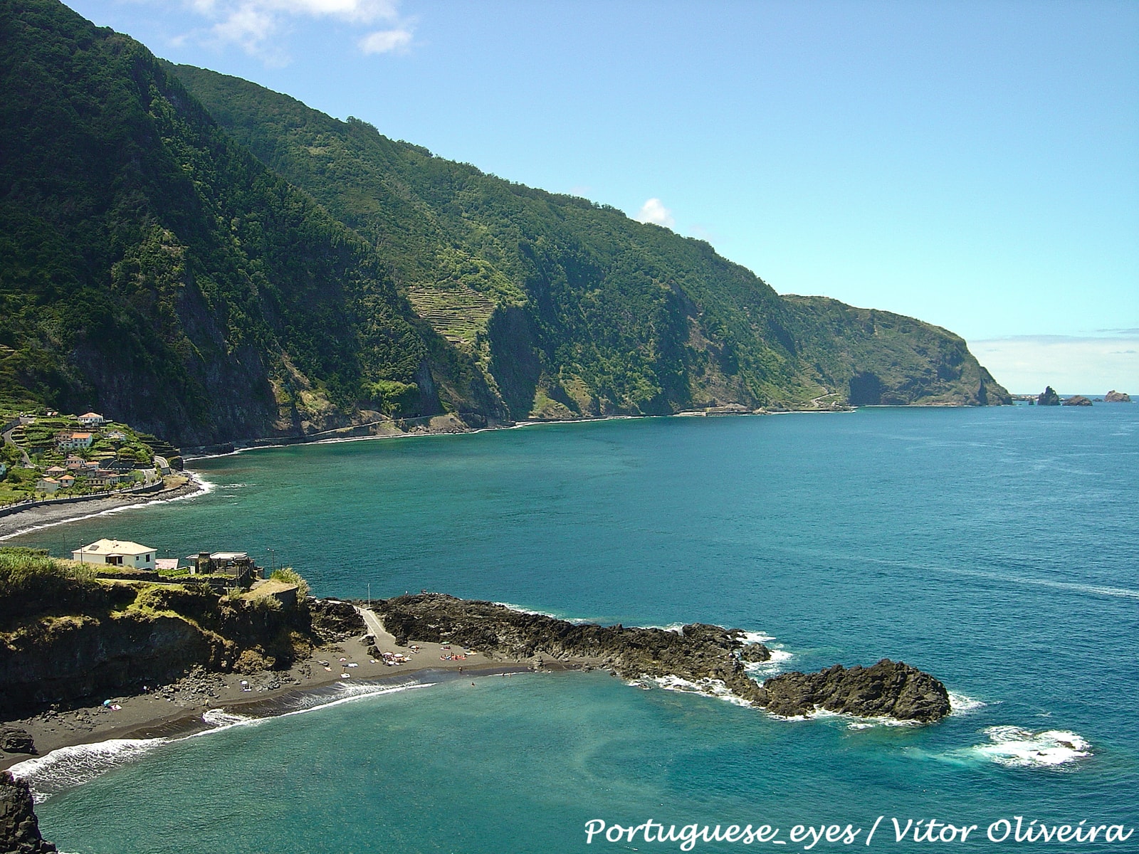 A dark sand or pebble beach with a dramatic sea stack just offshore and waves rolling in.