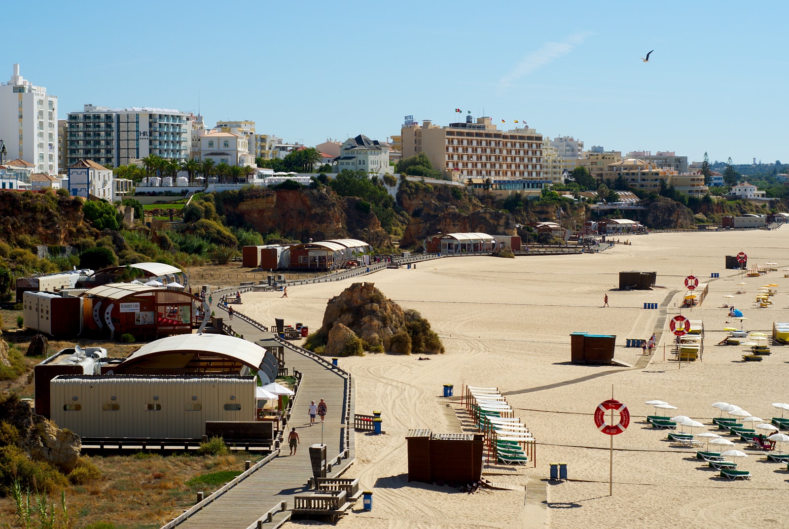 Beach kiosks, colorful umbrellas, and walkways on the sand at Praia da Rocha, with hotels visible atop the cliffs in the background.