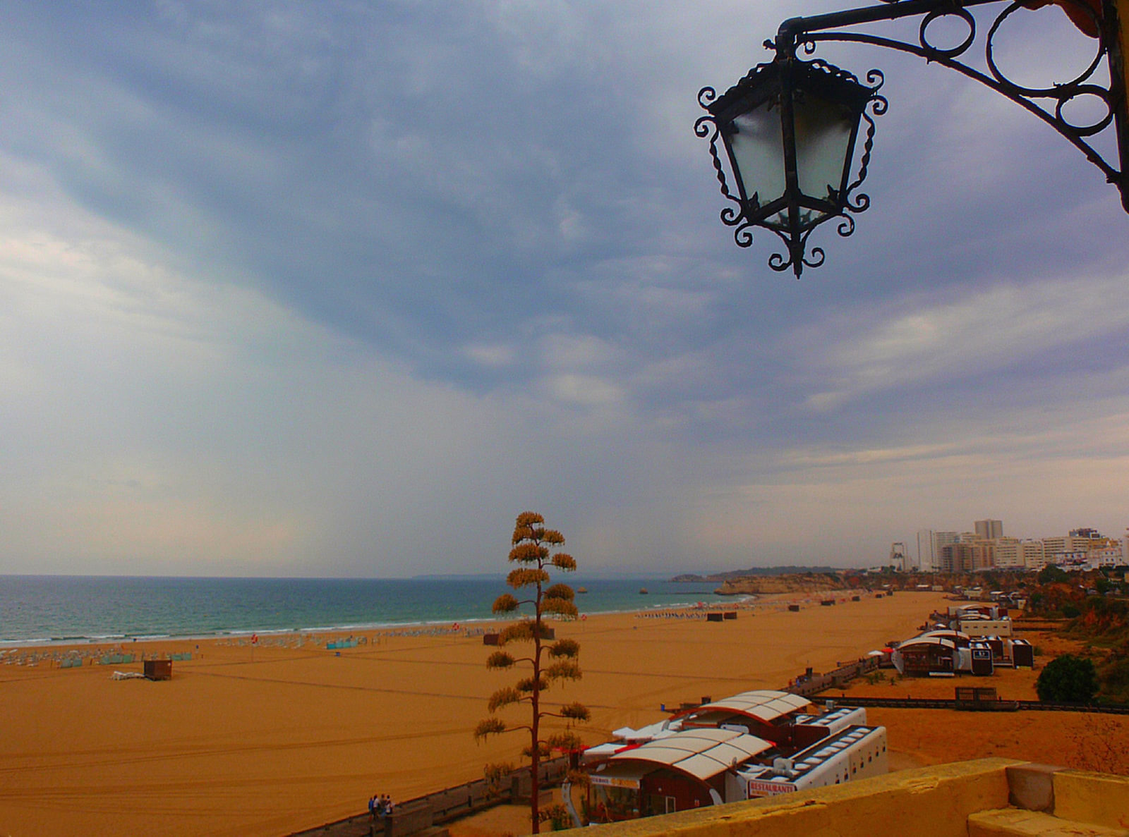 A view from a terrace overlooking Praia da Rocha, with a street lamp in the foreground and the beach and ocean in the background under a dramatic sky.