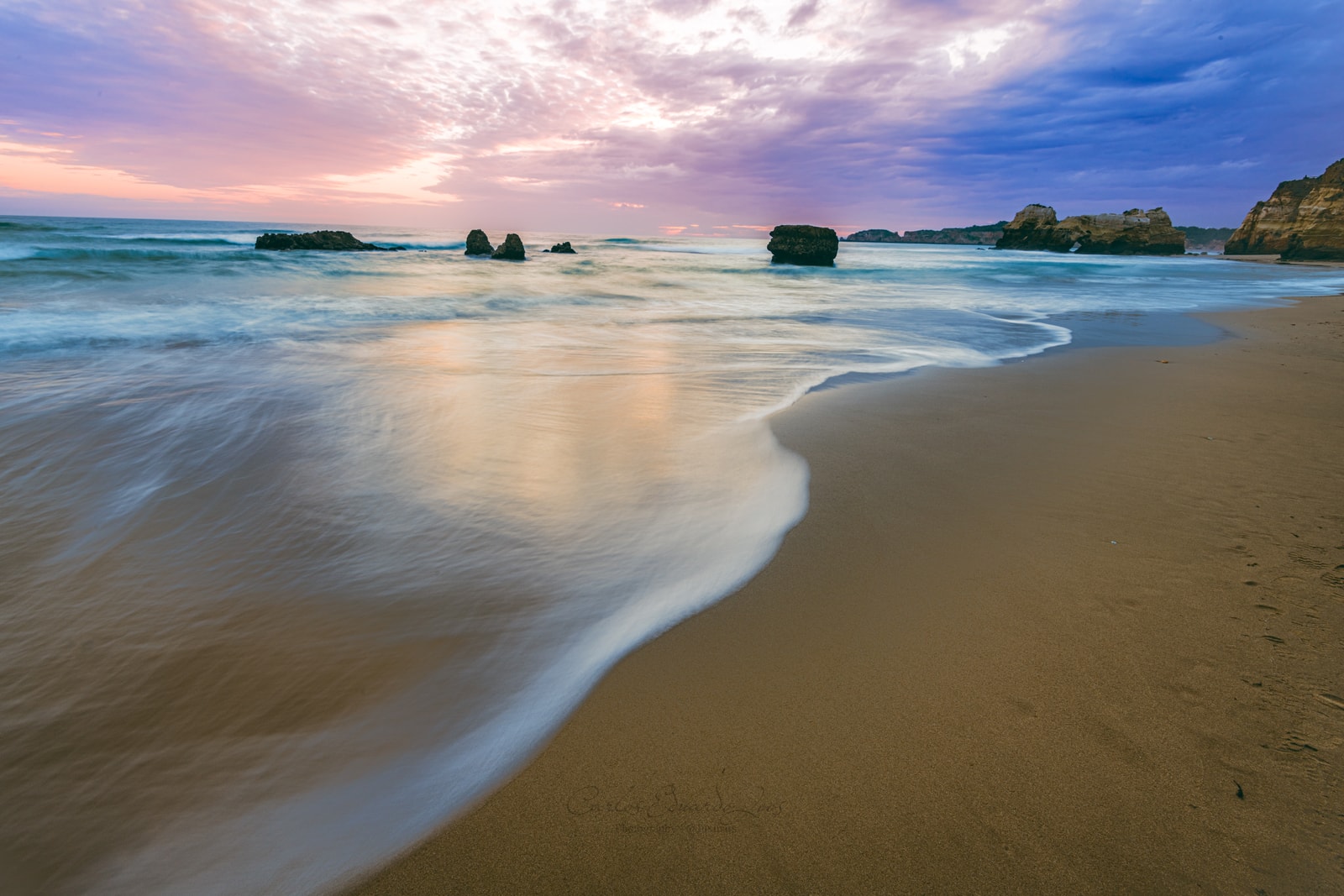 The sun sets over Praia da Rocha, casting a warm glow on the wet sand, the water, and the sea stacks along the coastline.