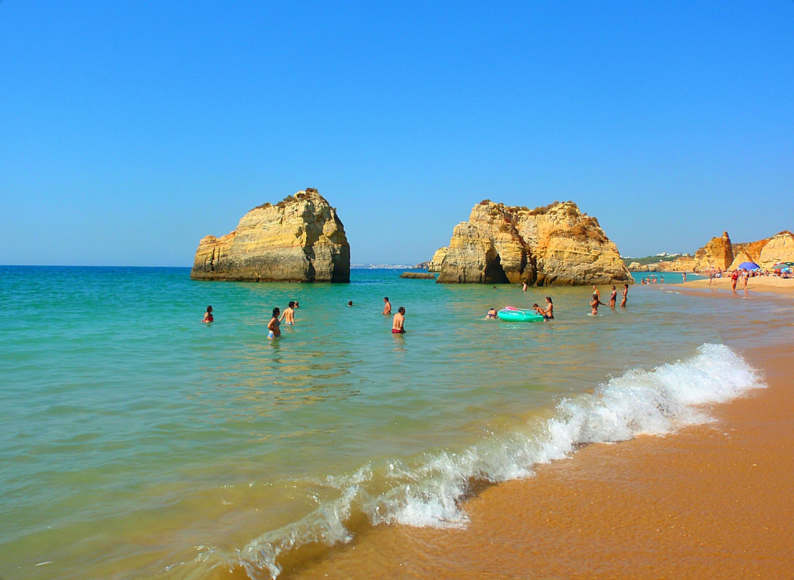 A breaking wave at Praia da Rocha with swimmers in the water and the beach's golden rock formations in the background.