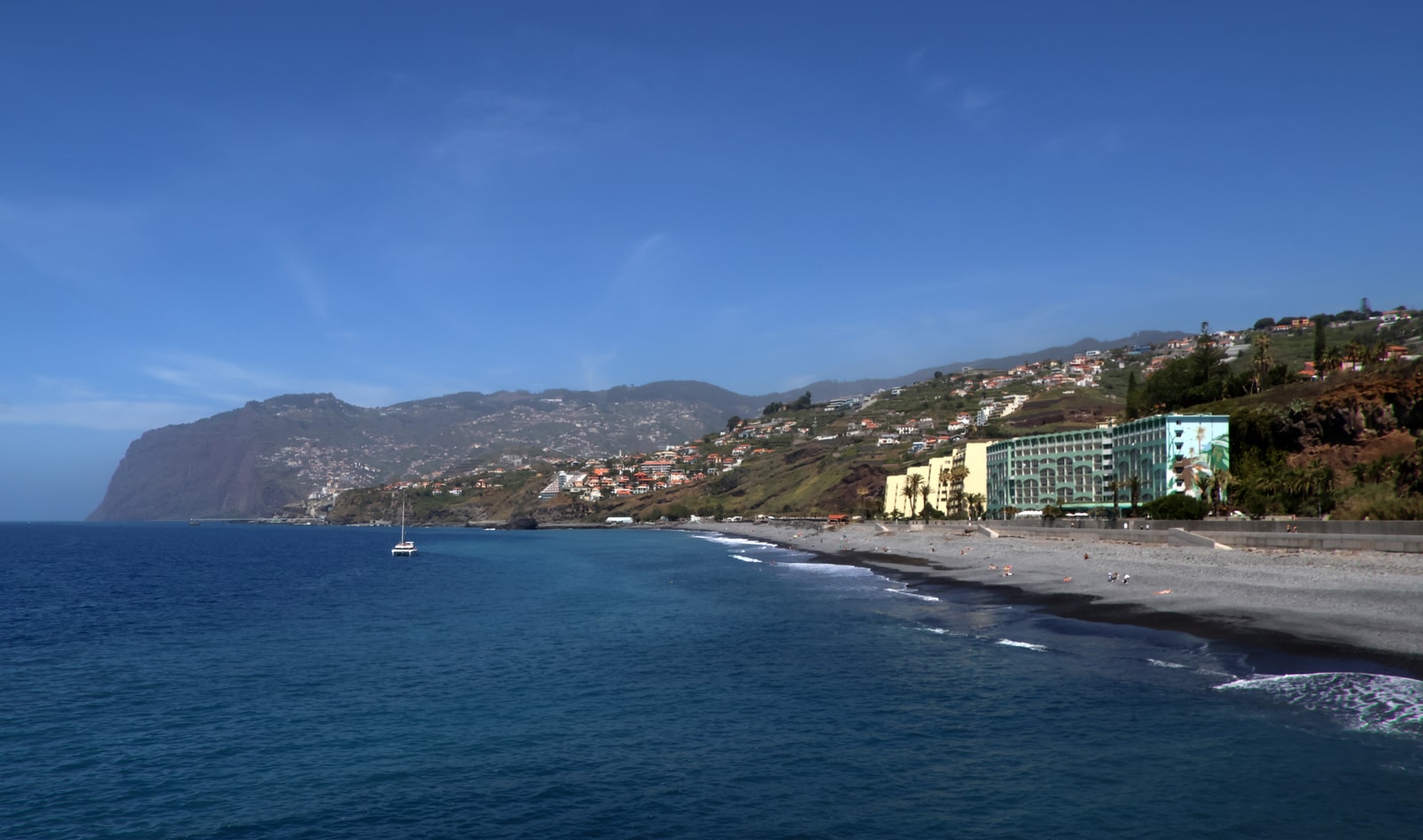 A sweeping view of a long pebble beach curving along a bay, with hills and buildings rising in the background under a partly cloudy sky.