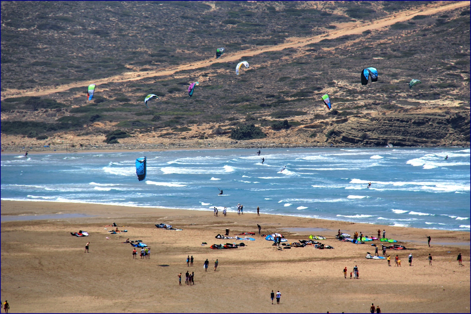 Kitesurfers and windsurfers are scattered across a wide, sandy beach with their colorful equipment.