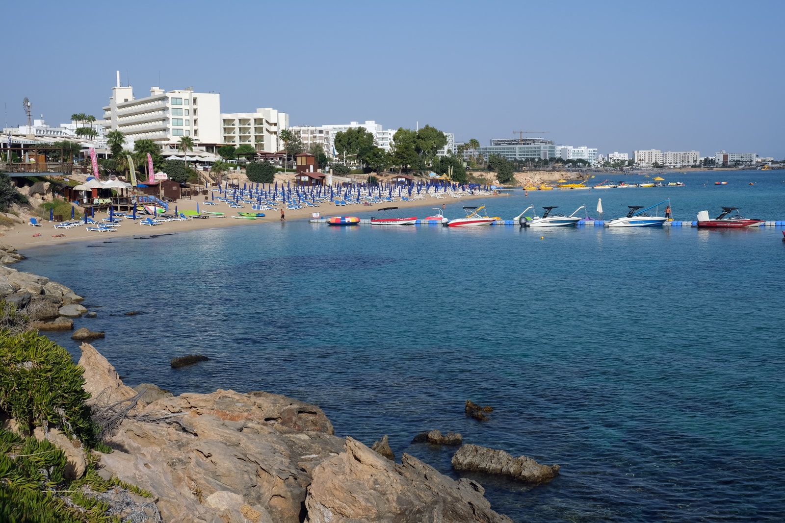 A view of Protaras Beach showing a rocky foreground with the sandy beach, umbrellas, and resort hotels in the distance.