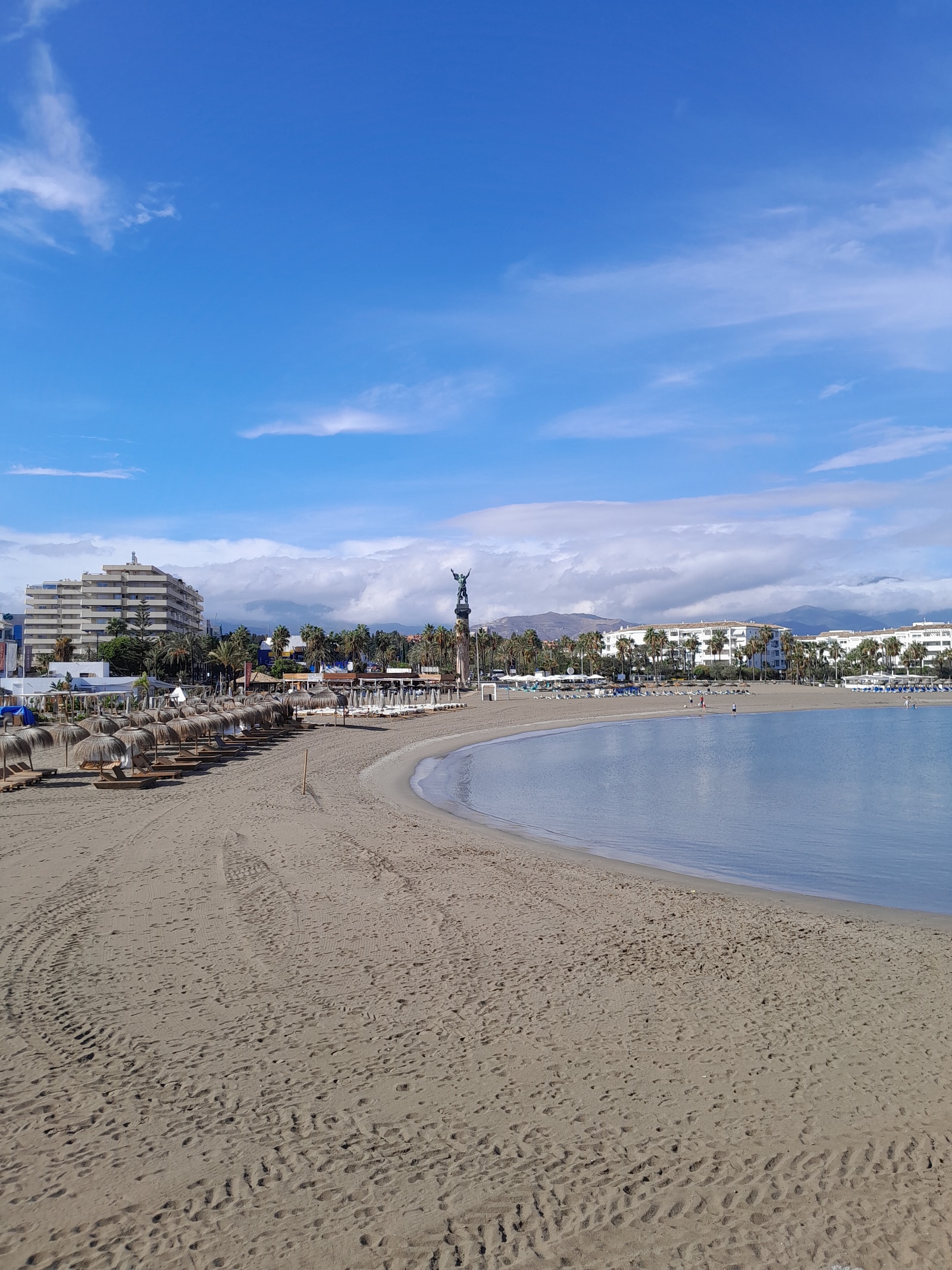 A portrait view of the wide, sandy Puerto Banús beach with rows of straw umbrellas and a prominent waterfront monument column in the background.