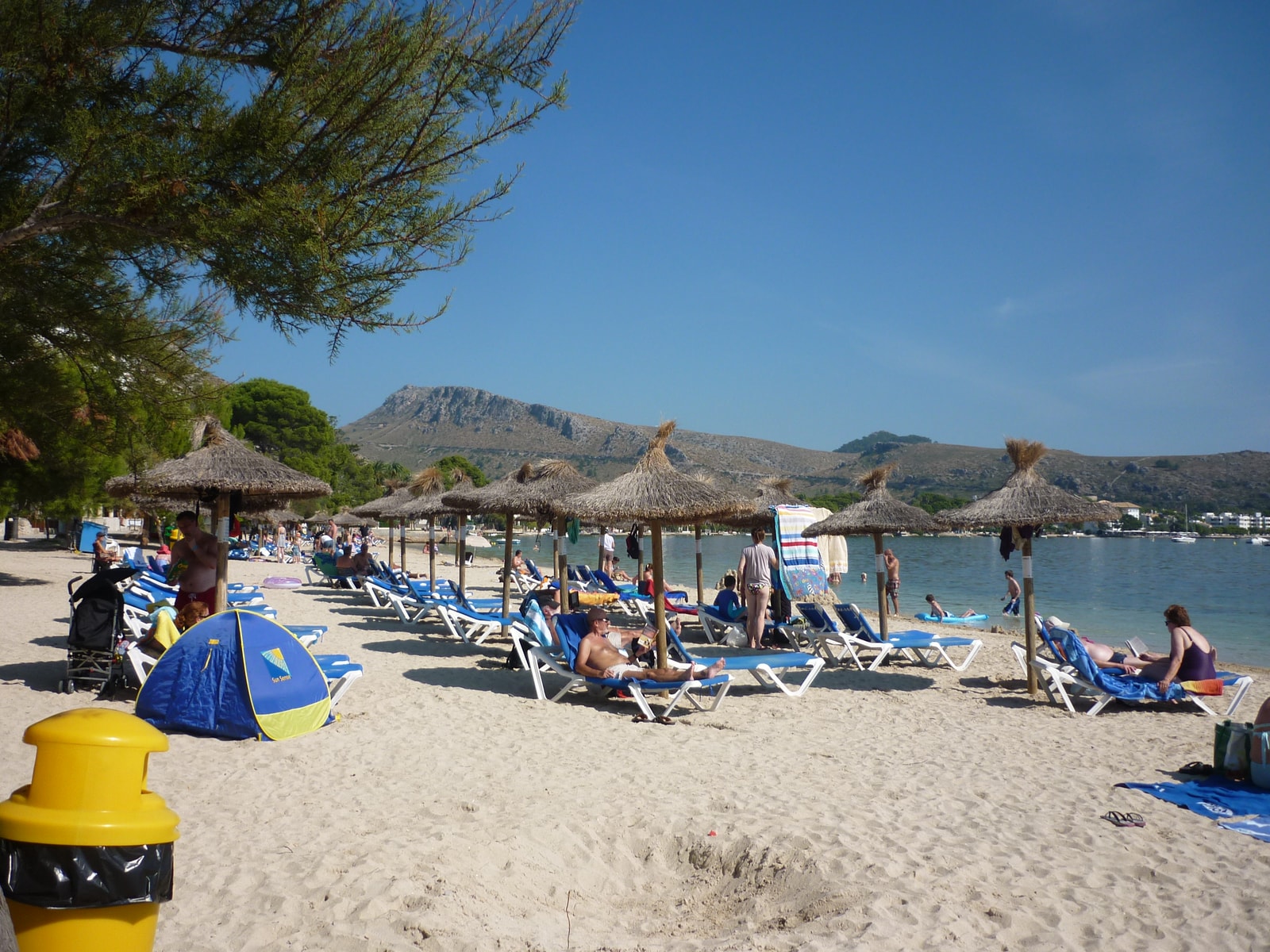 A wide sandy beach with blue and white umbrellas, sun loungers, and people swimming in calm water with mountains in the distance.