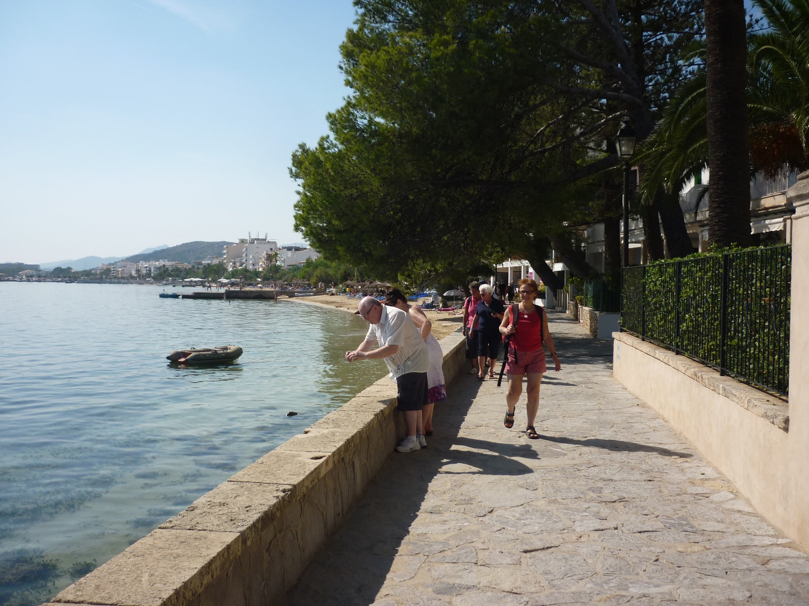 A stone-paved promenade runs along the water's edge, shaded by large pine trees, with a small boat in the calm blue sea.