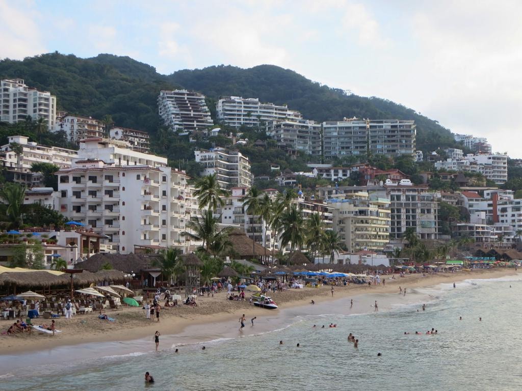A bustling Puerto Vallarta beach with sunbathers and umbrellas, backed by hotels and apartments built into the green hillside.