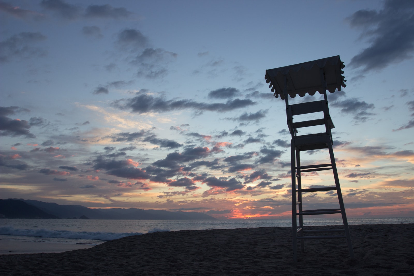 A silhouetted lifeguard chair on a Puerto Vallarta beach faces a beautiful orange and purple sunset over the ocean and mountains.