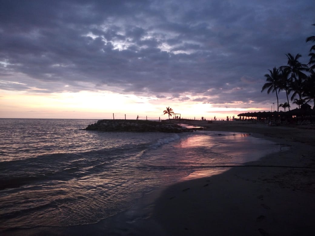 A dramatic sunset with orange clouds reflects on the wet sand of a Puerto Vallarta beach near a rocky jetty.