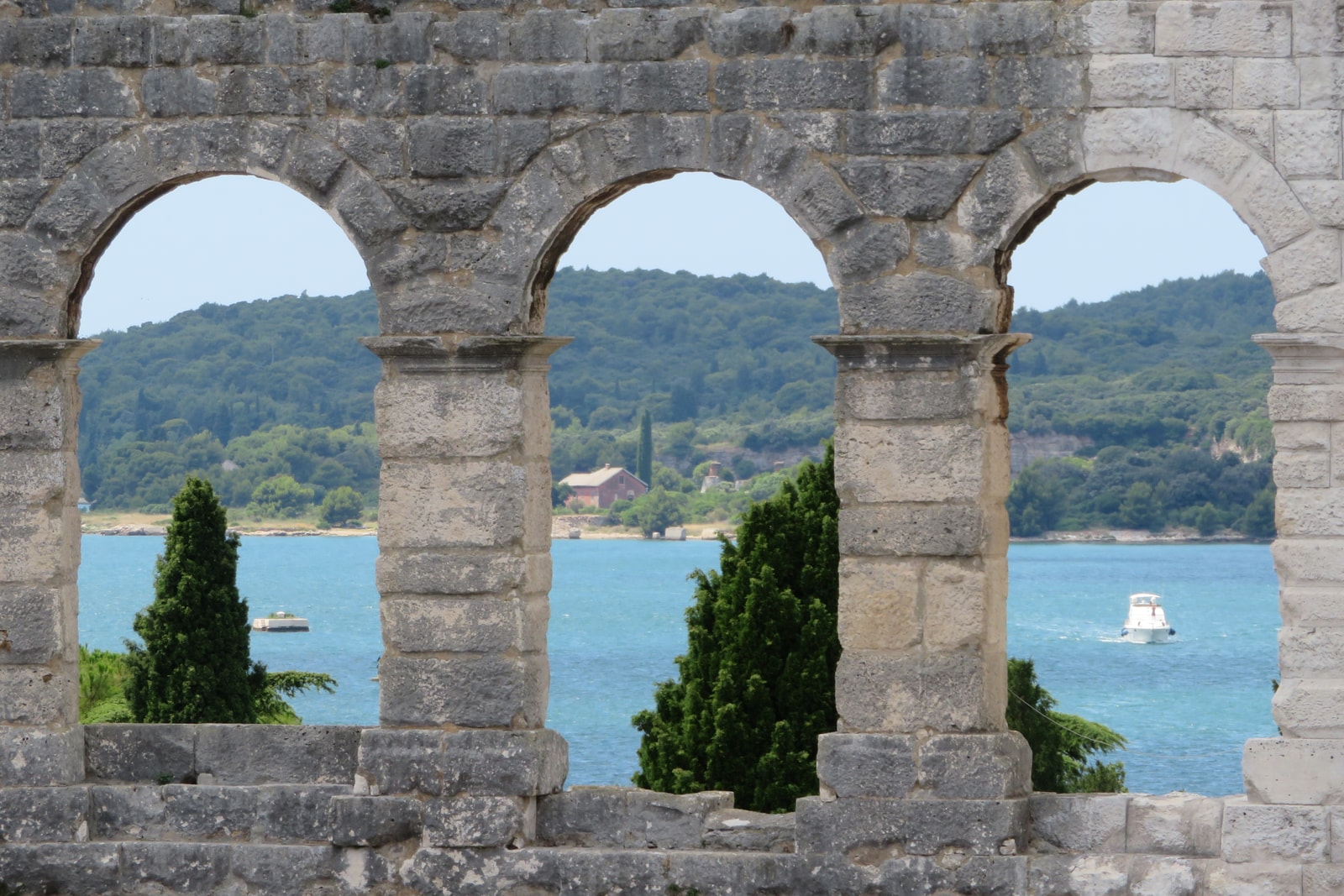 The ancient Roman Arena in Pula, with its limestone arches framing a view of the turquoise Adriatic Sea and pine-covered coastline.