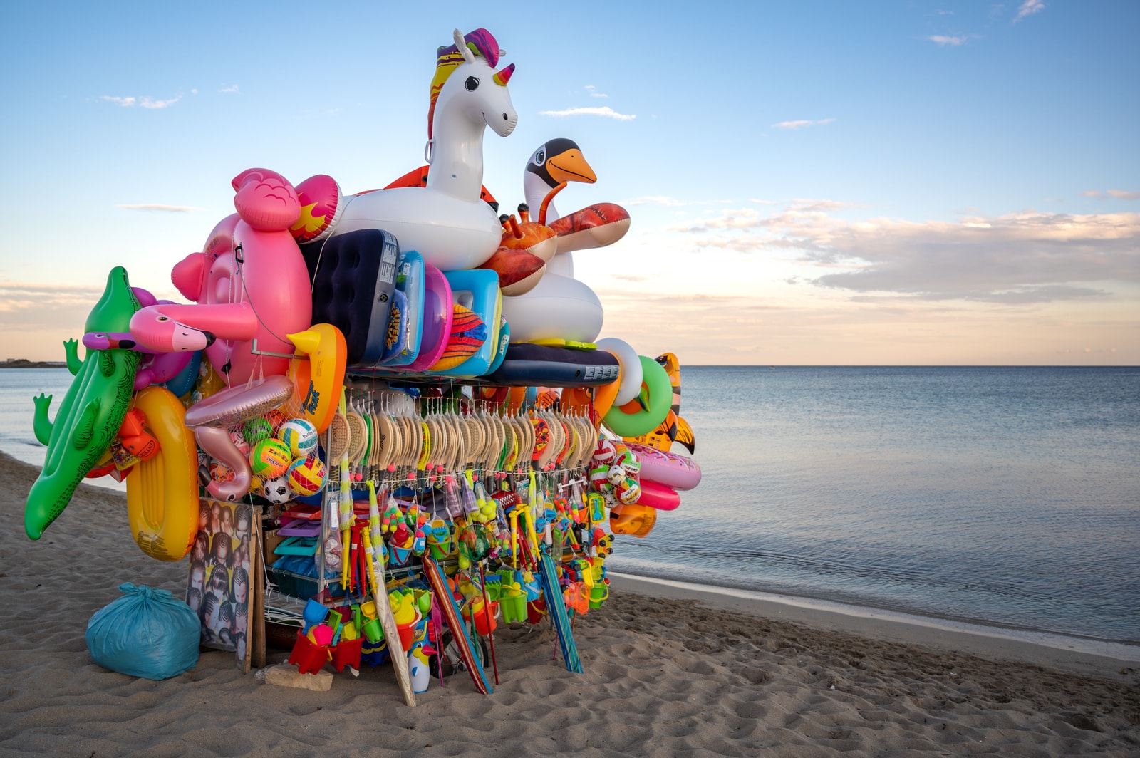 Colorful beach inflatables, including swans and unicorns, stand on the sand at Punta Prosciutto, with the sunset sea in the background.