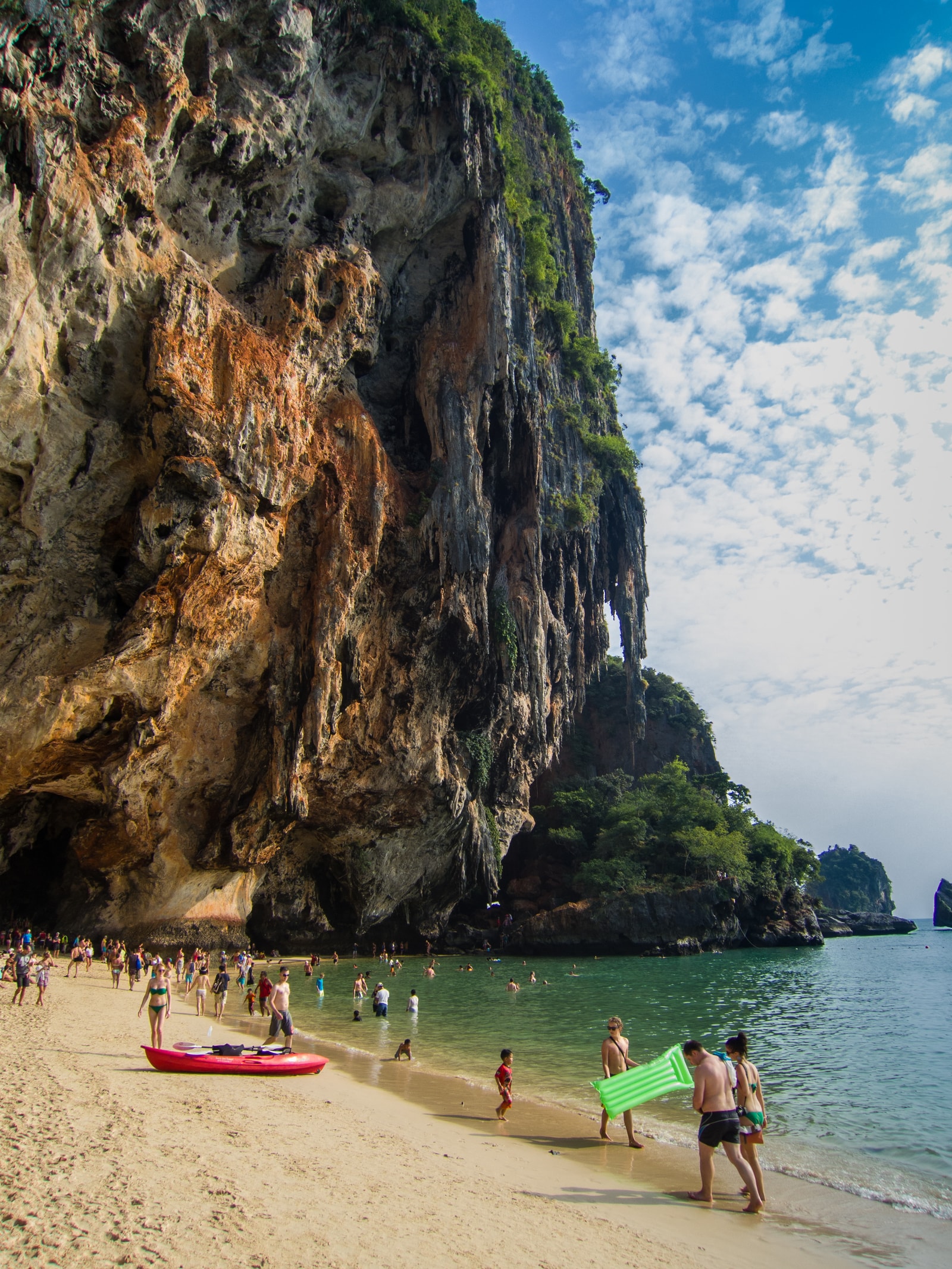 People swim and kayak in the clear turquoise water at the base of a towering limestone cliff at Railay Beach.