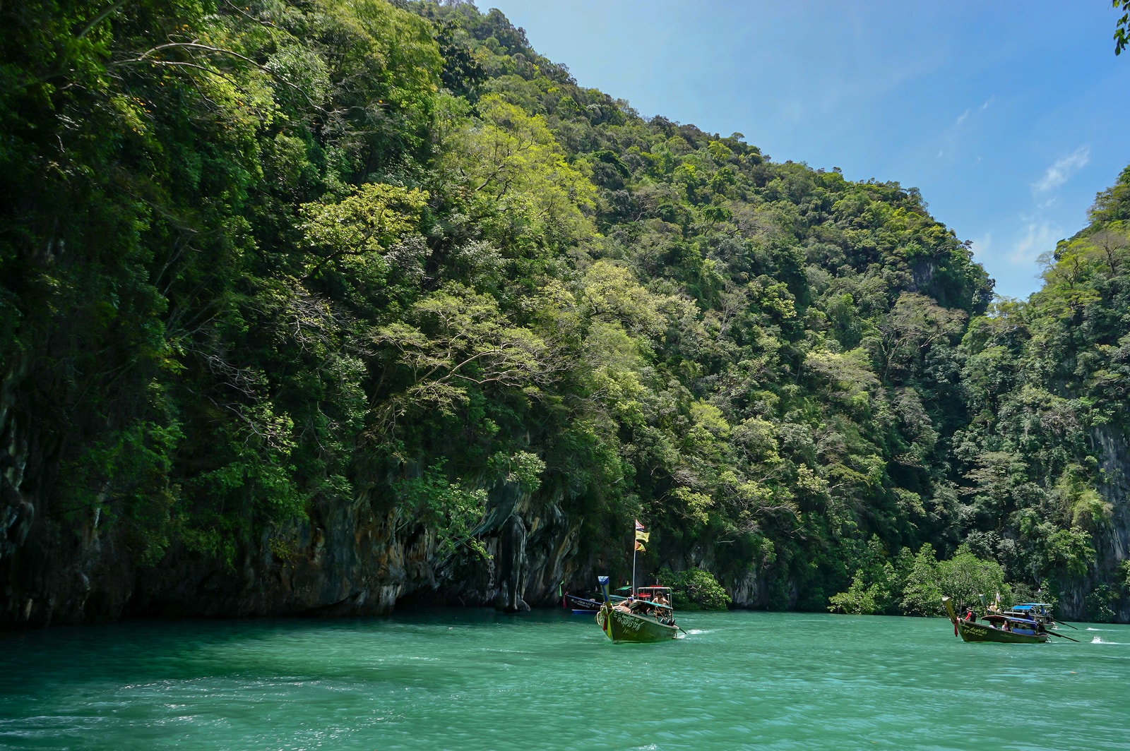 A lone longtail boat motors through a tranquil inlet surrounded by towering, jungle-covered limestone karsts under a bright sky.