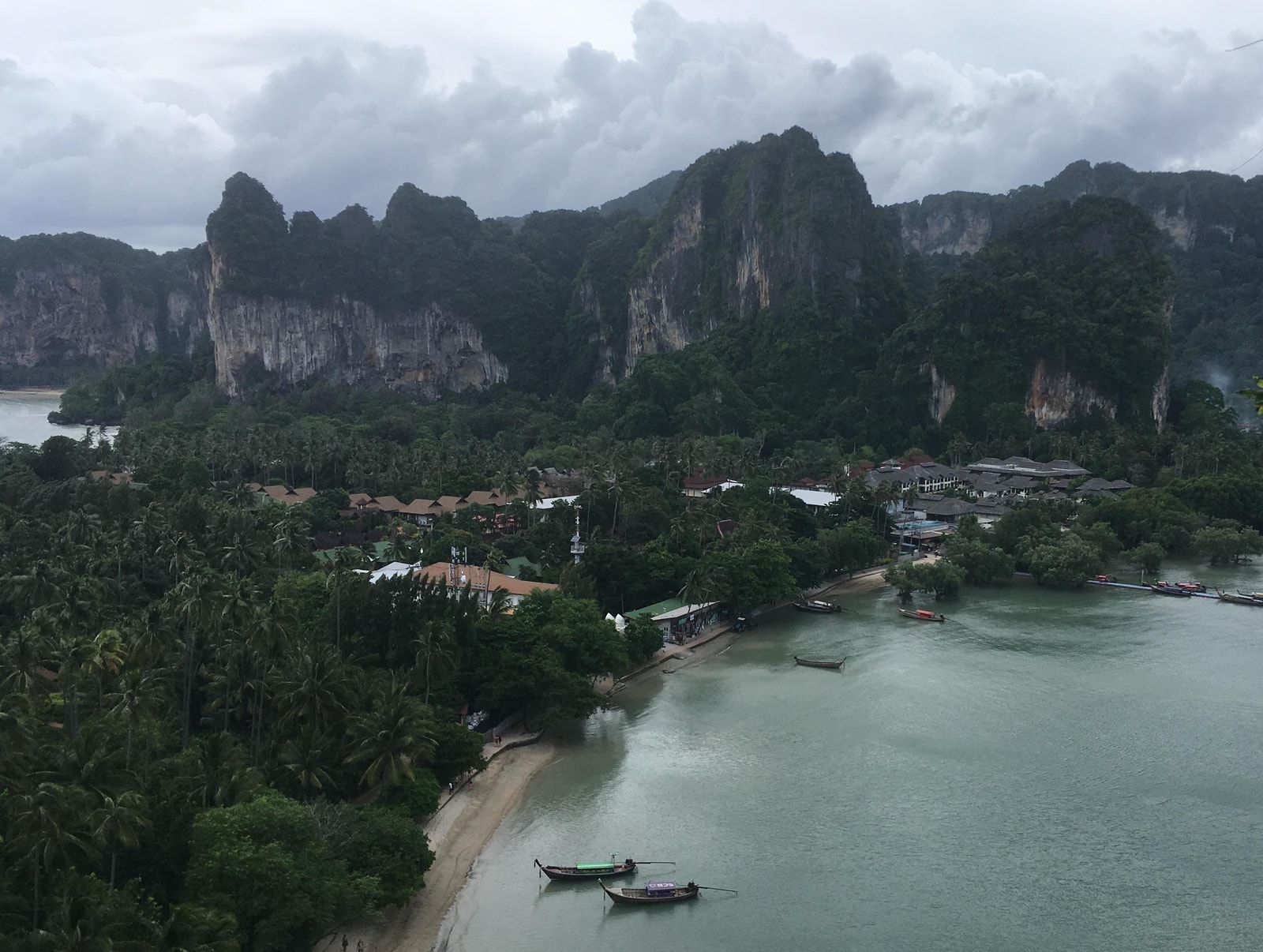 An elevated, panoramic view from the Railay viewpoint shows the peninsula's two bays, with the narrow strip of land covered in green vegetation and dotted with buildings, framed by dramatic limestone cliffs.