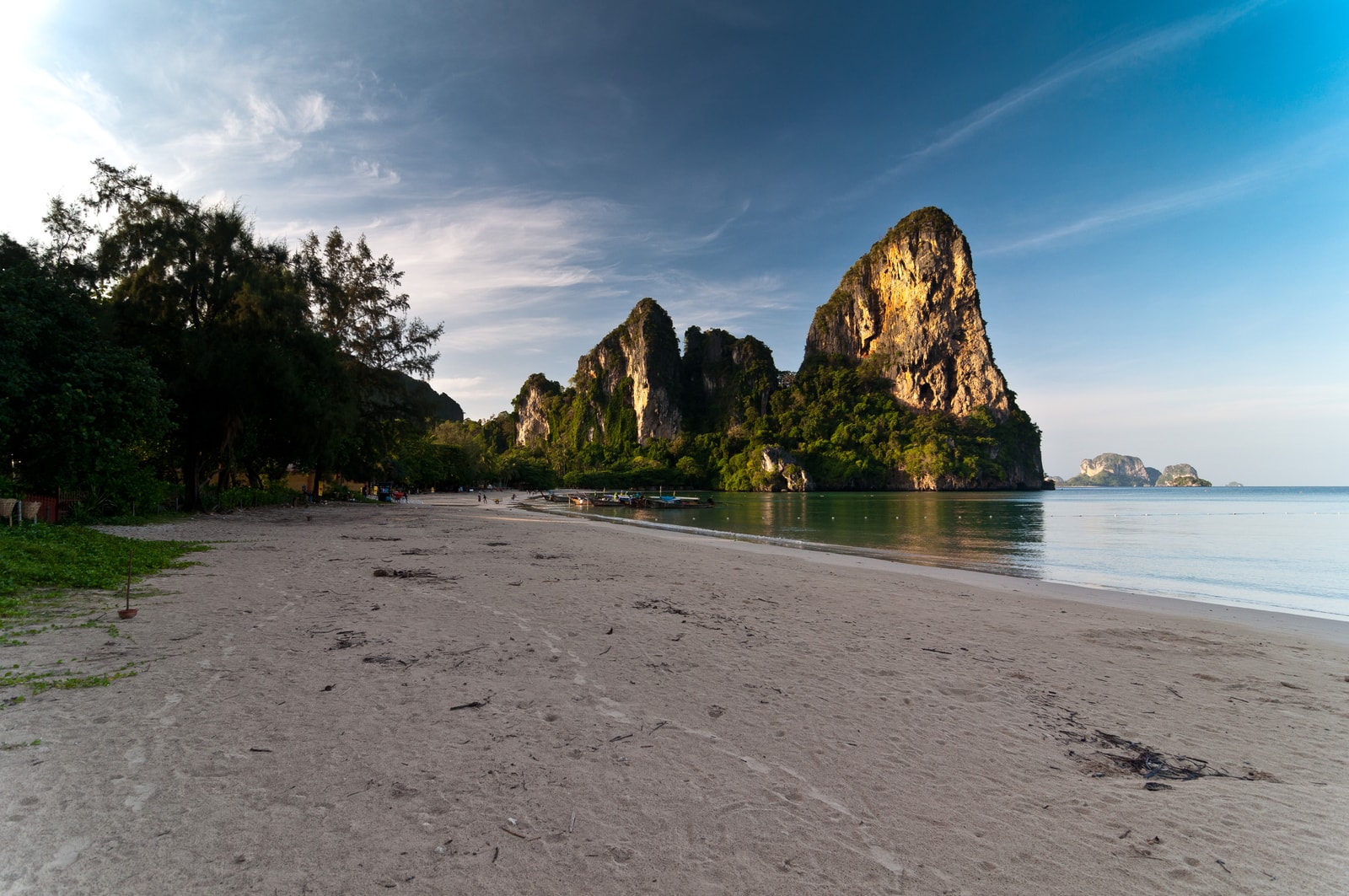 The sun rises over Railay Beach, casting a warm golden light on the distant karst formations, while the sand remains in cool shadow.