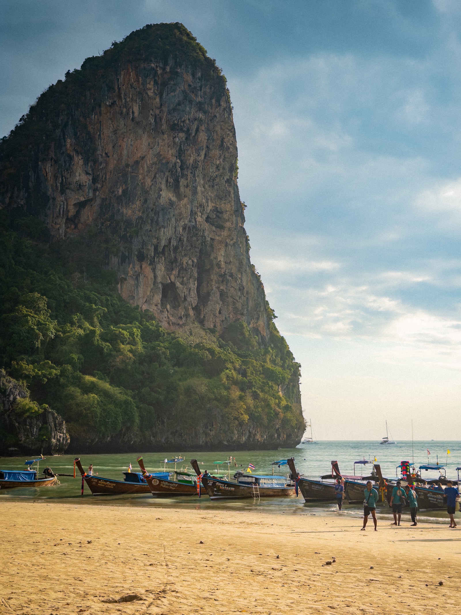 A vertical shot shows a massive, sheer limestone cliff towering over longtail boats parked on the sandy shore of Railay West.