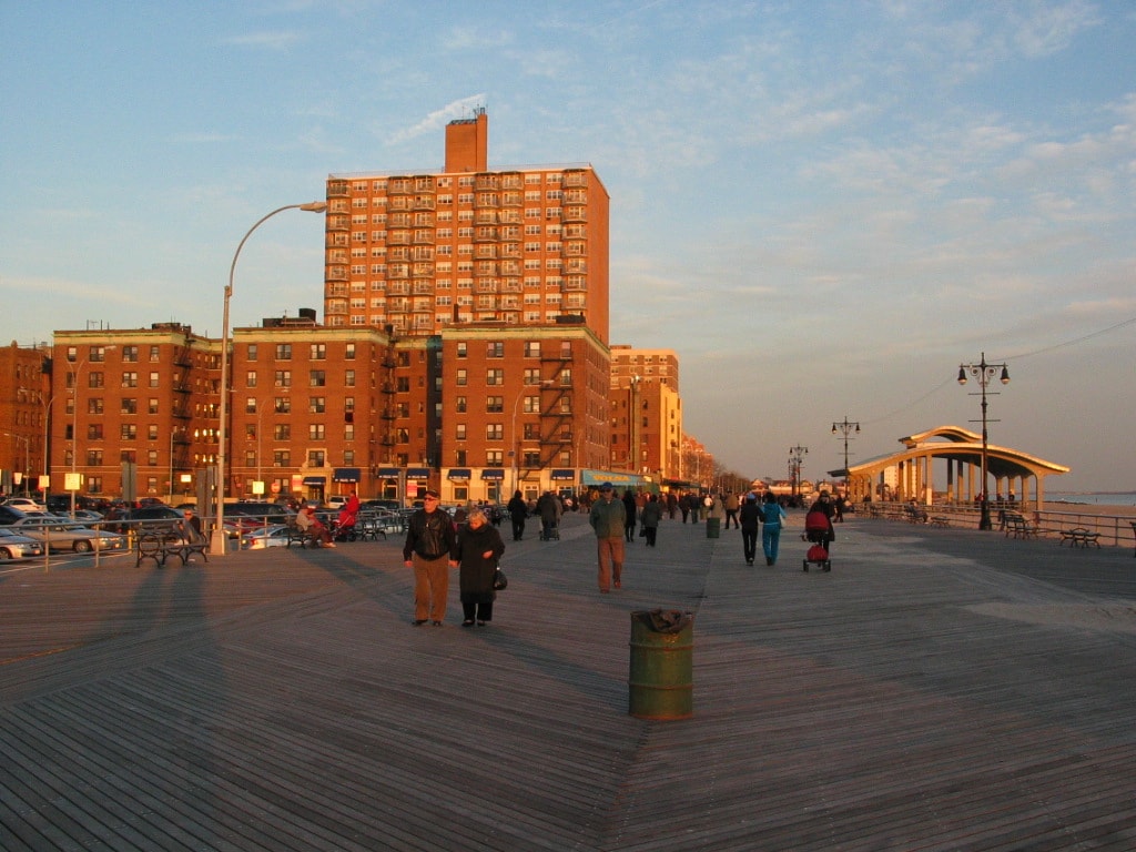 The wooden planks of the Riegelmann Boardwalk at sunset, with residential apartment buildings lining the background.